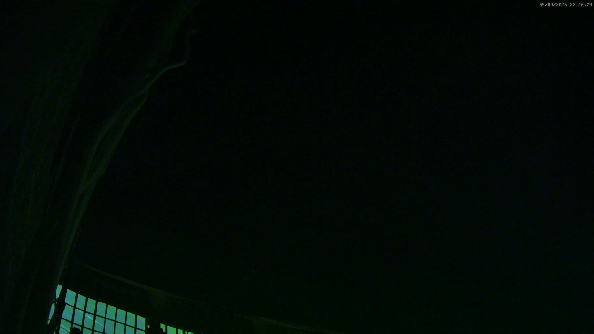 A low-angle, nighttime view shows a dark sky and a portion of a building with a gridded window illuminated from within.