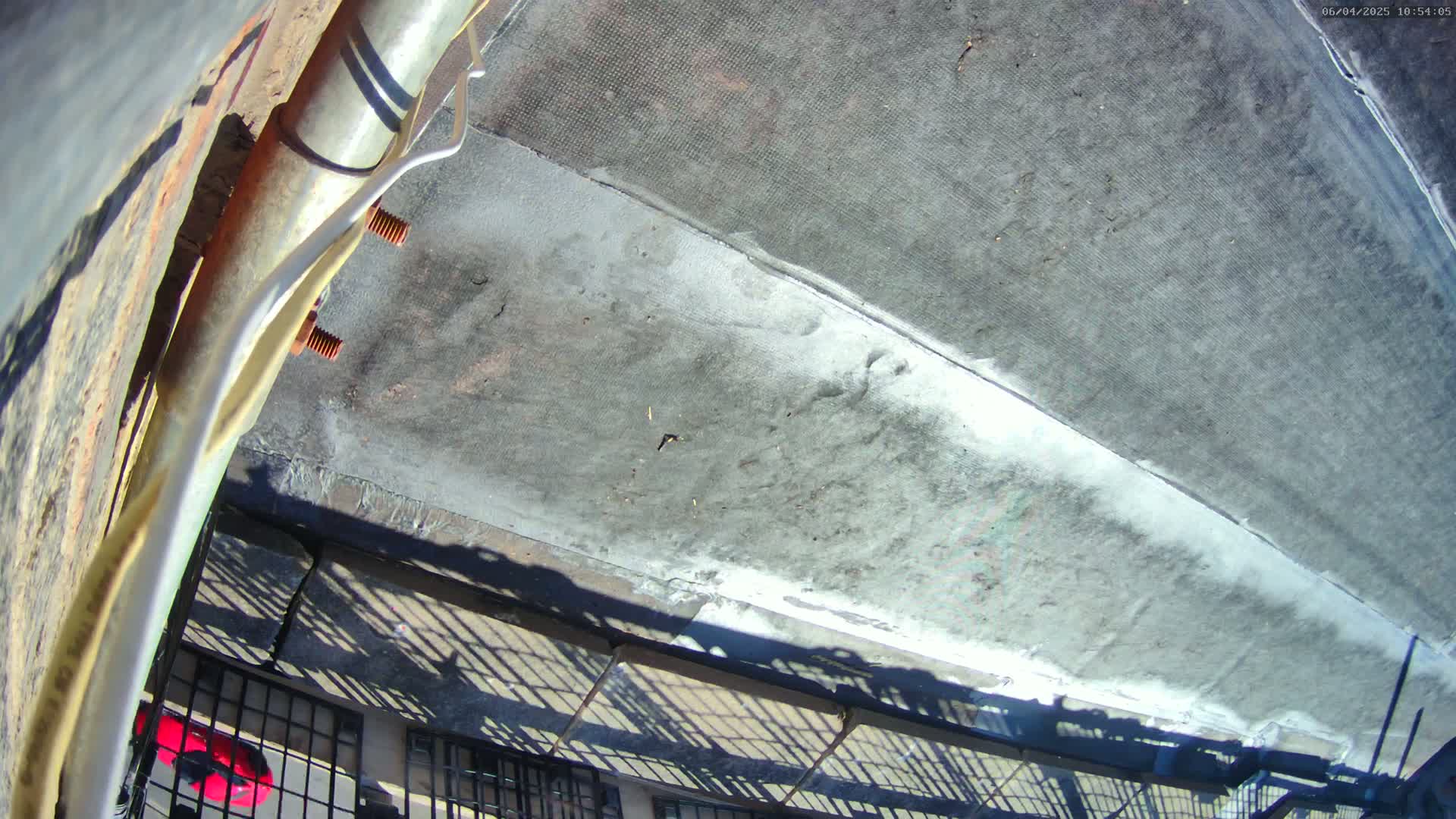 A high-angle, outdoor view shows a flat, gray rooftop with a section of lighter-colored patching, partially visible rusty metal, and a metal railing casting shadows in the sunlight.