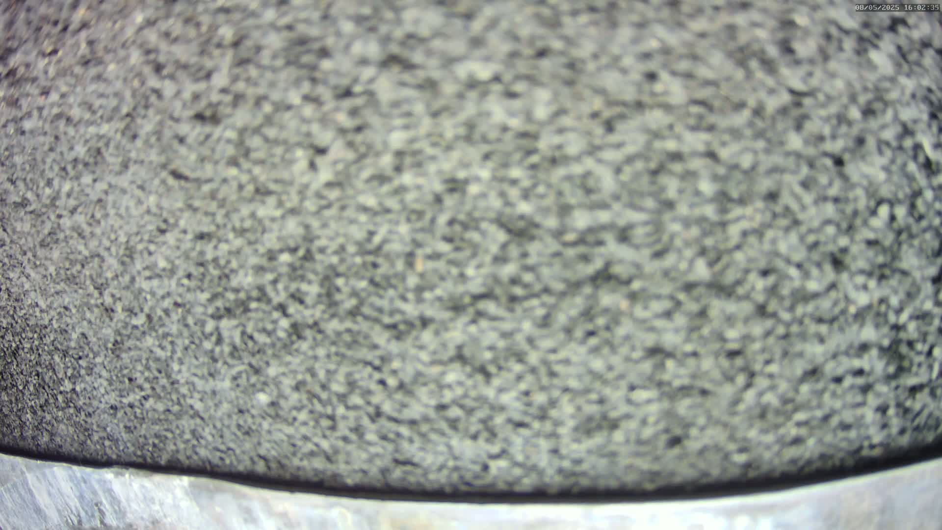 A close-up view shows a blurry, dark gray, granular surface that appears to be asphalt or similar material, with a slightly lighter gray metal edge at the bottom.