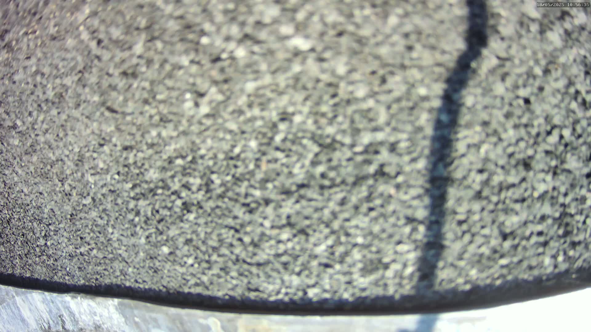 A close-up view of a dark gray, pebbly surface with a long shadow cast across it on a sunny day.