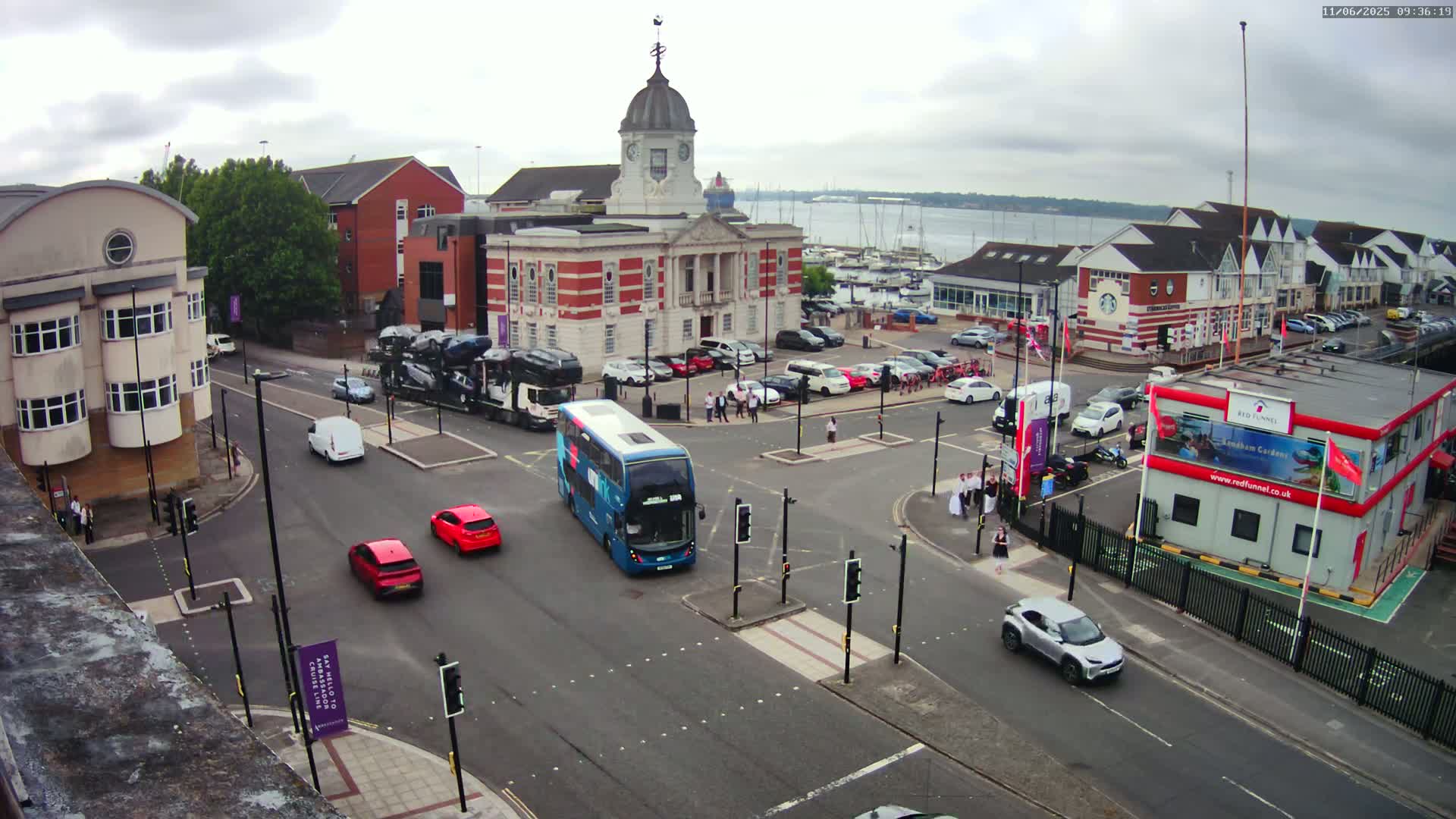 A high-angle, overcast-day view of a town square with a large clock tower building, several cars, a double-decker bus, and people walking near a marina.