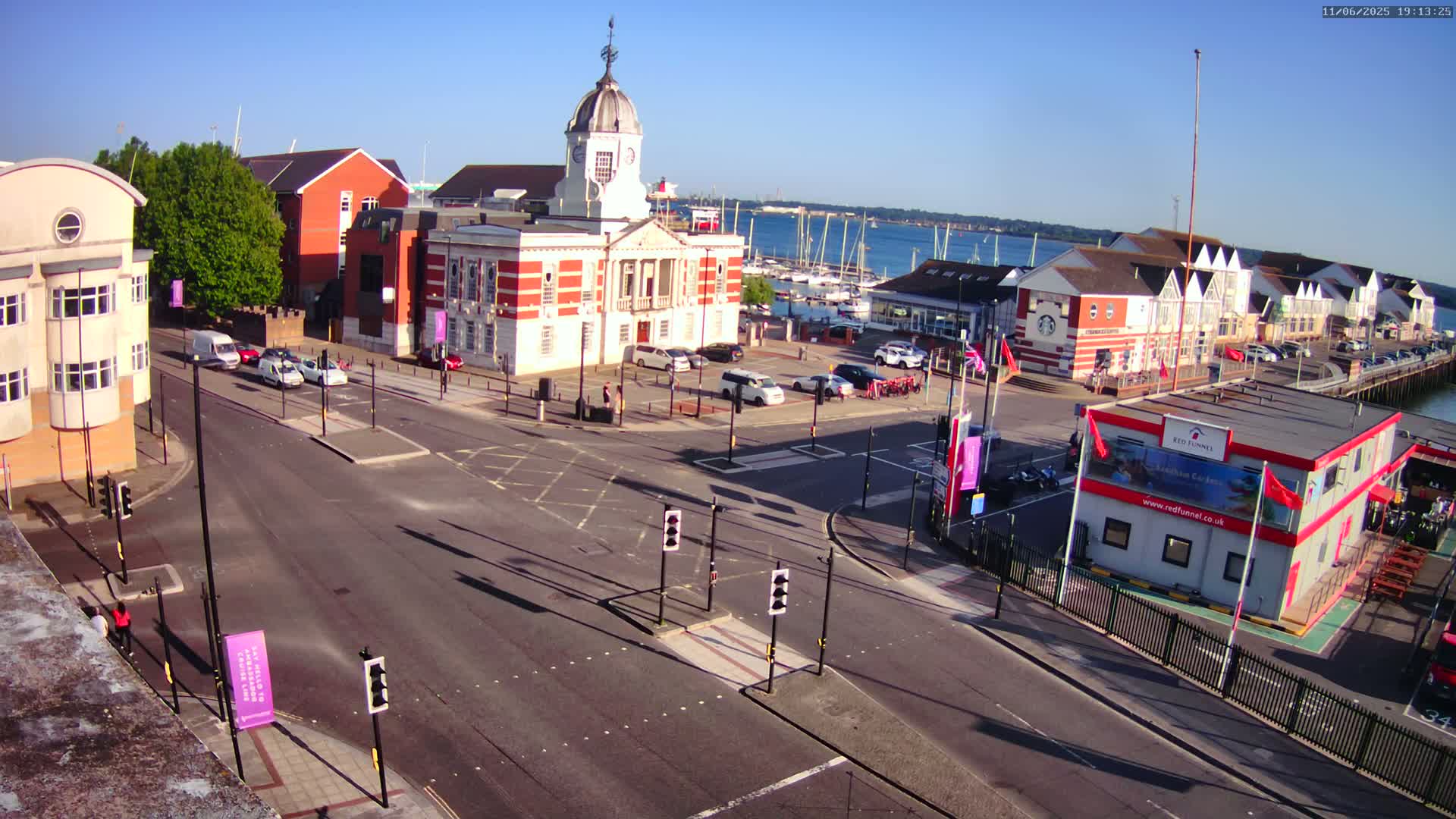 A sunny day overlooking a town square with a red-brick building, roads, and parked cars near a marina with boats under a clear sky.
