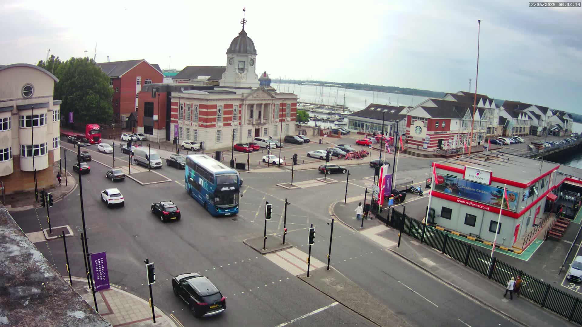 A view of a town square with a clock tower, several buildings, and cars and buses driving on intersecting roads under an overcast sky.