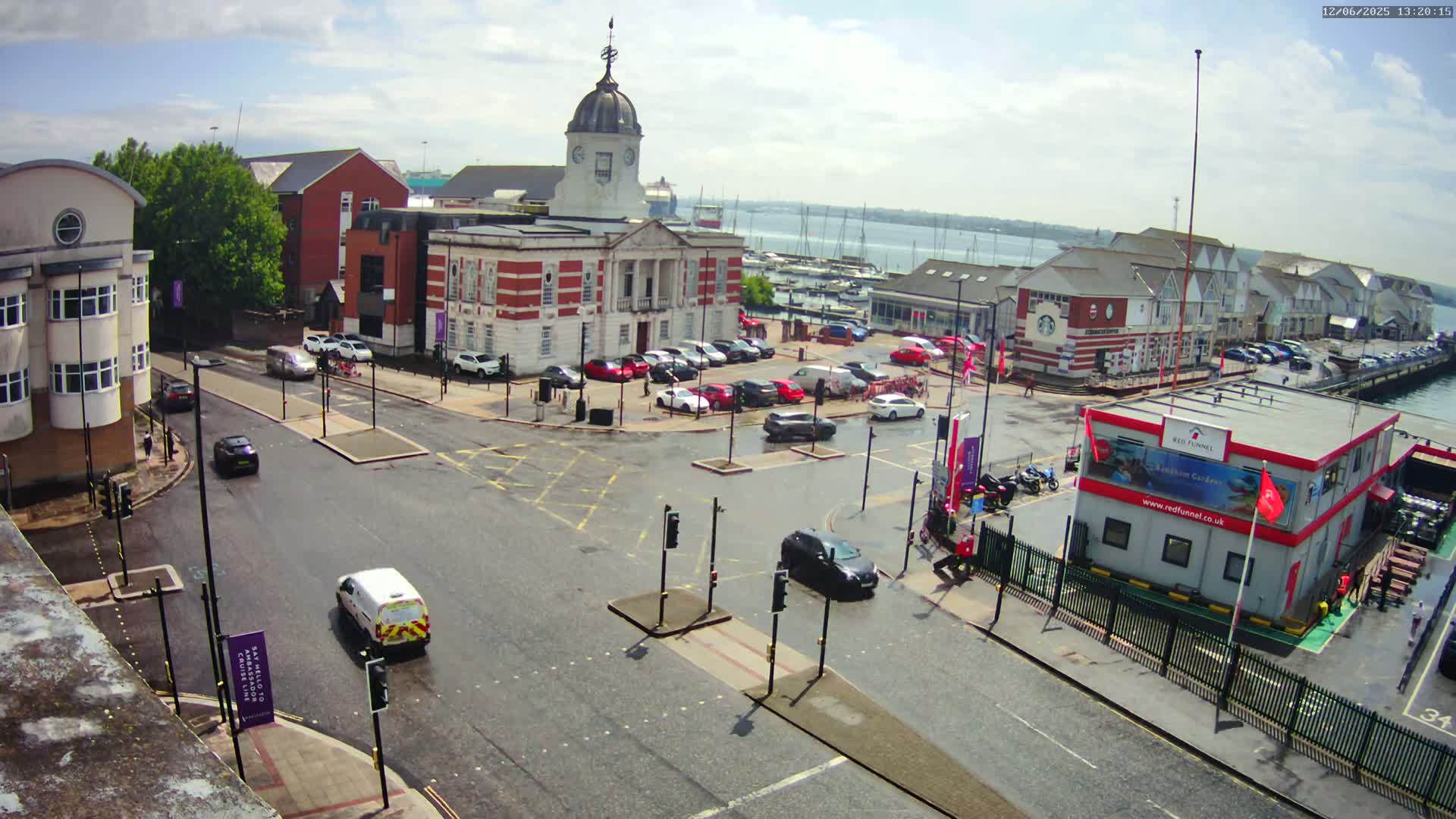A wet, partly sunny day shows a town square with a brick building and a parking lot,  surrounded by other buildings, with a harbor and boats visible in the background.