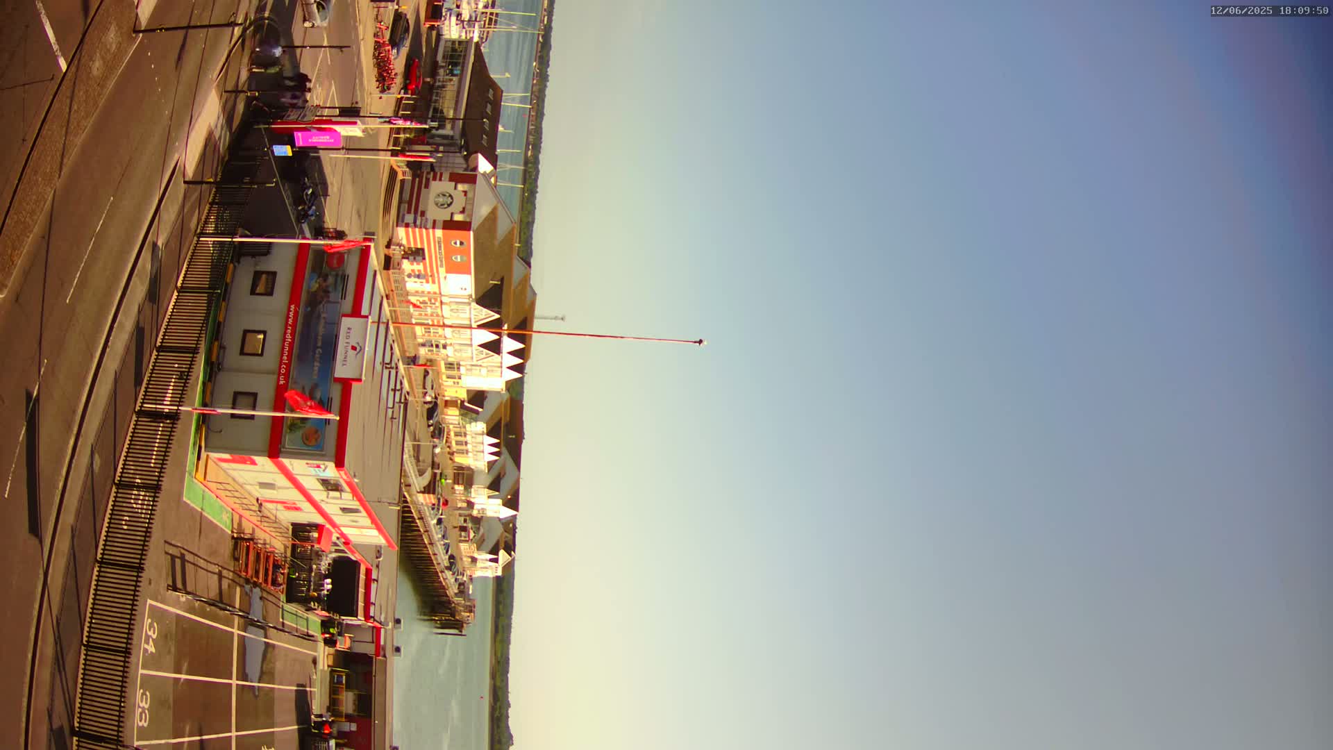 A high-angle view of a waterfront area on a clear, sunny day shows buildings, a pier, a road, and a parking lot.