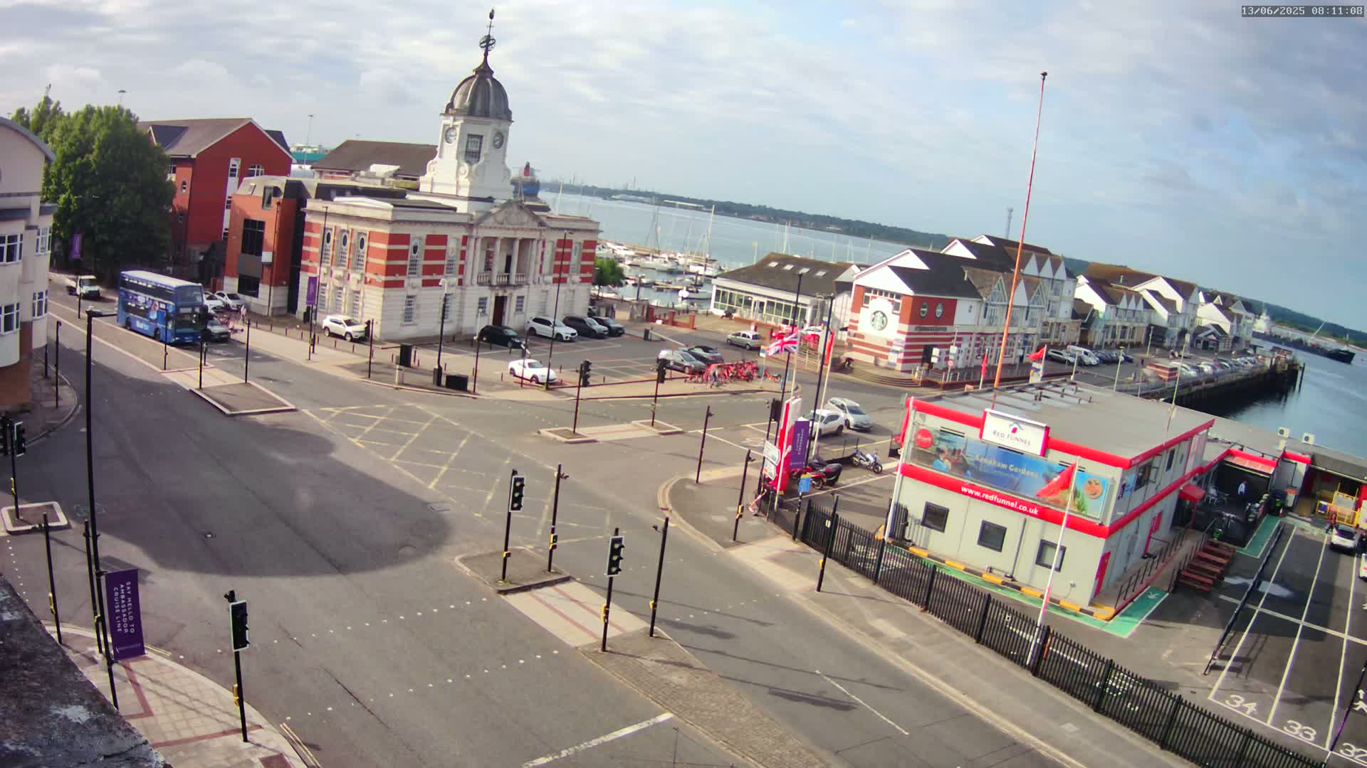 A partly sunny day overlooking a town square with a brick building, a parking lot, roadways, and a marina in the background.