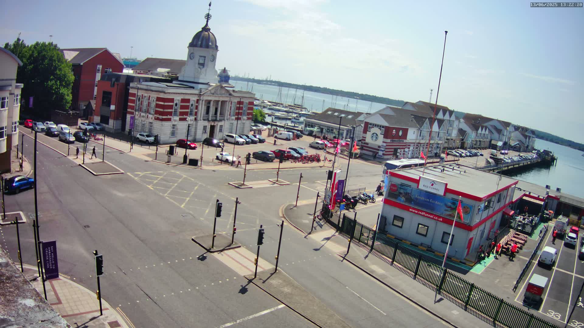 A sunny, high-angle view of a town square near a harbor, showing a brick clock tower, parked cars, pedestrian crossings, and a ferry terminal.