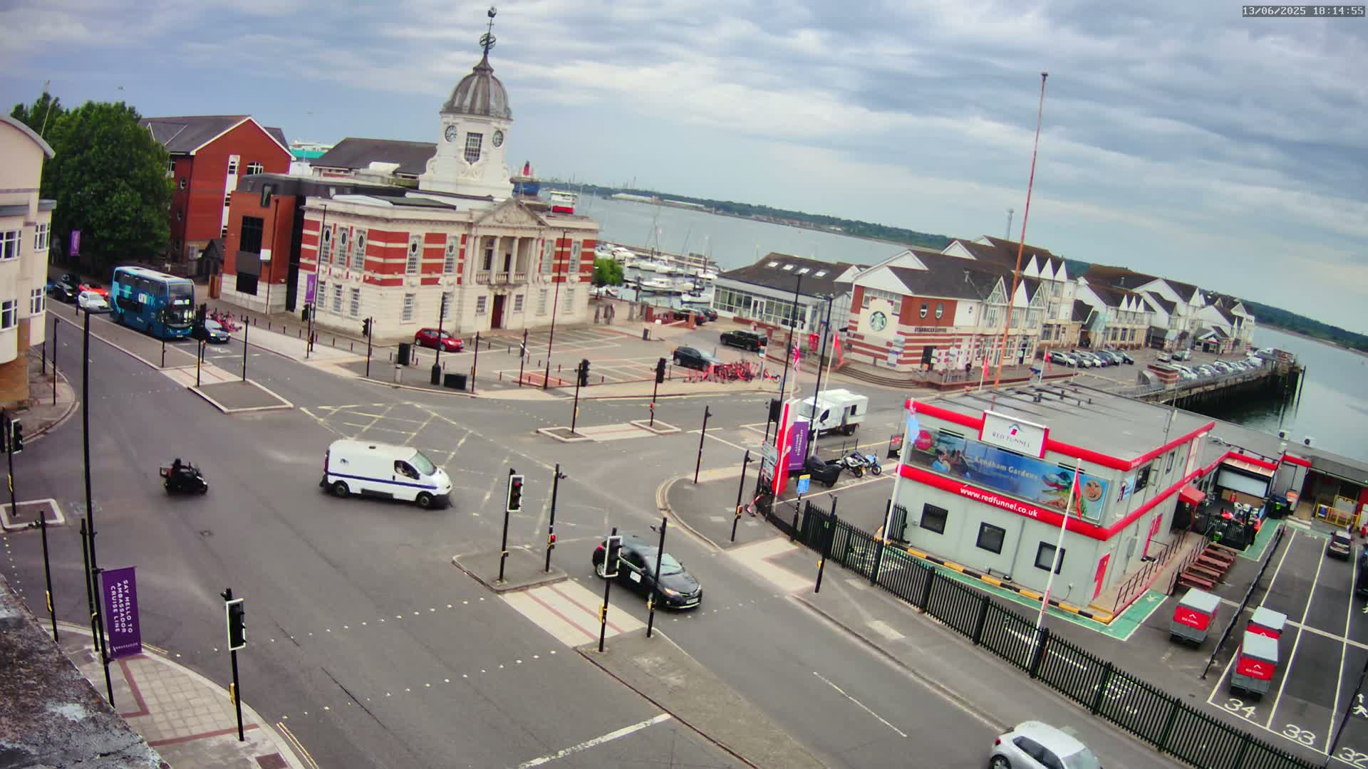 A high-angle view of a town square near a harbor on an overcast day, showing several vehicles at a busy intersection with a large brick building in the background.
