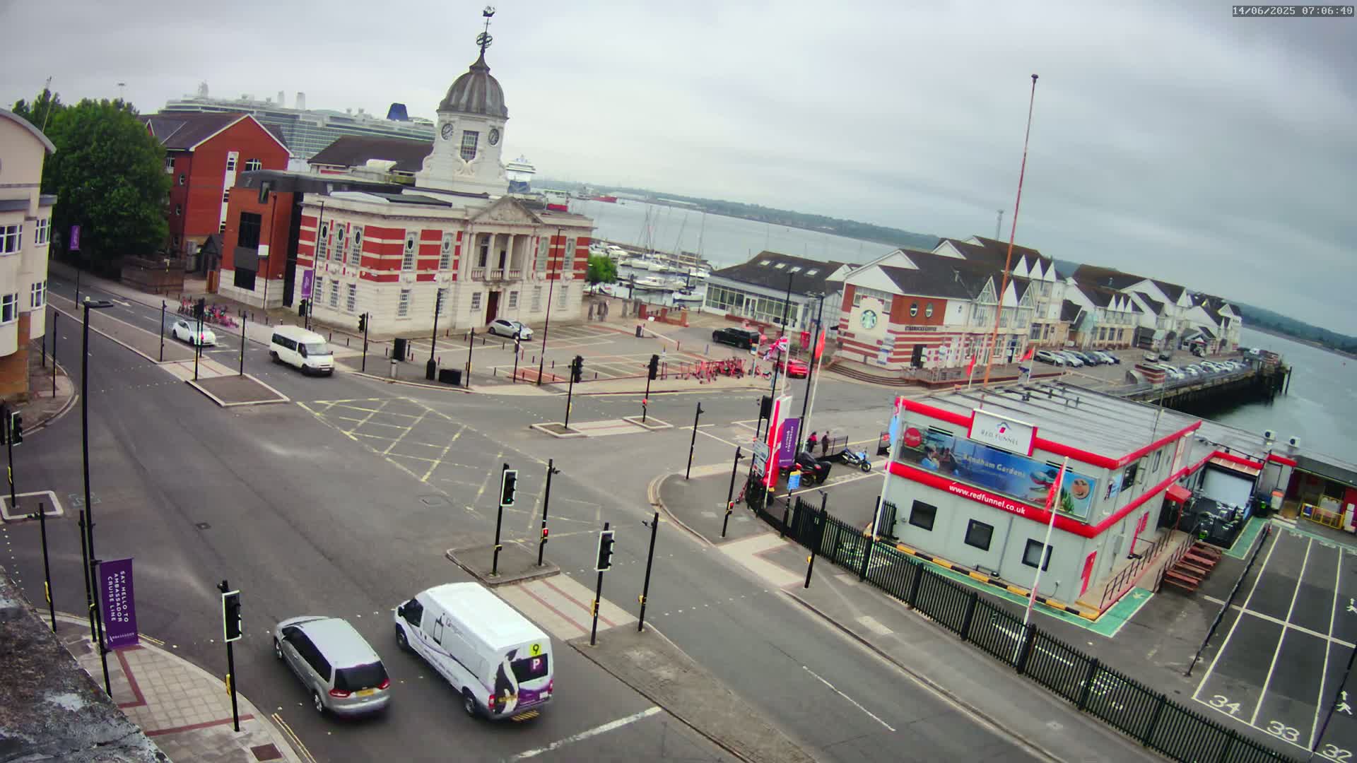 A high-angle view of a town square near a harbor on an overcast day, showing several vehicles at a busy intersection with a large brick building in the background.