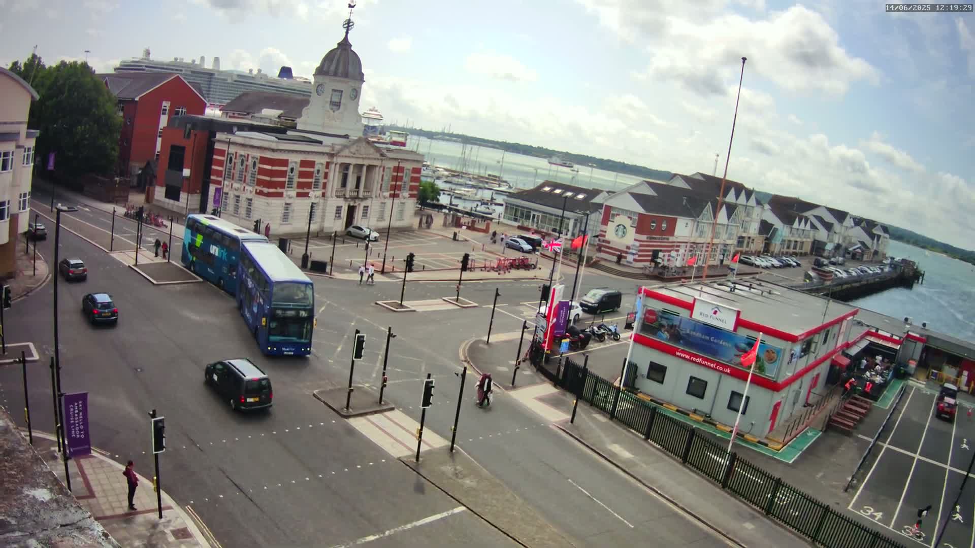 A partly cloudy day shows a street intersection with a double-decker bus, several cars, and pedestrians, near a waterfront with buildings and a large cruise ship in the distance.