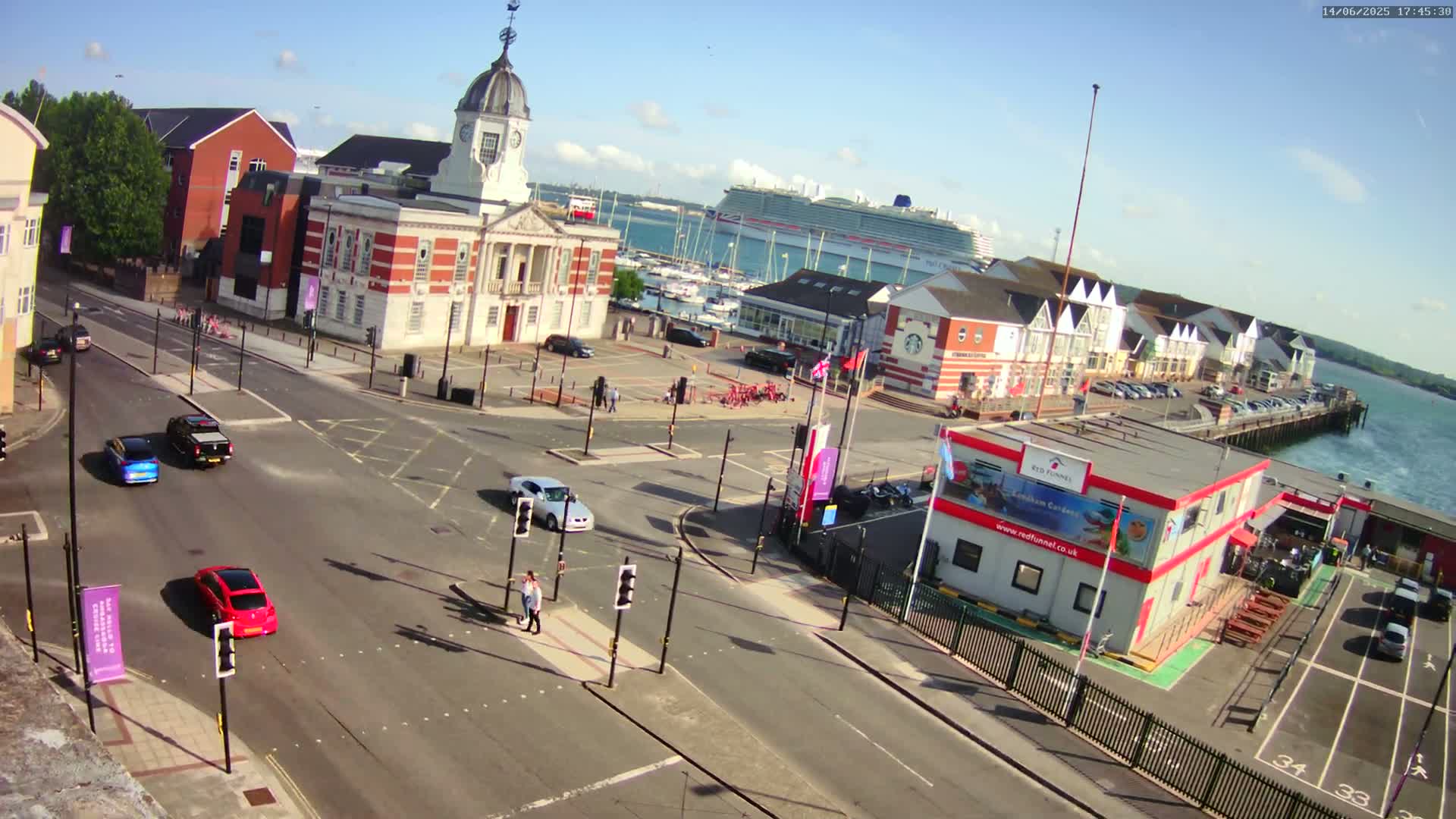 A sunny harbor scene shows a town square with several cars at an intersection, a large building with a clock tower, a cruise ship in the distance, and a waterfront with docked boats.