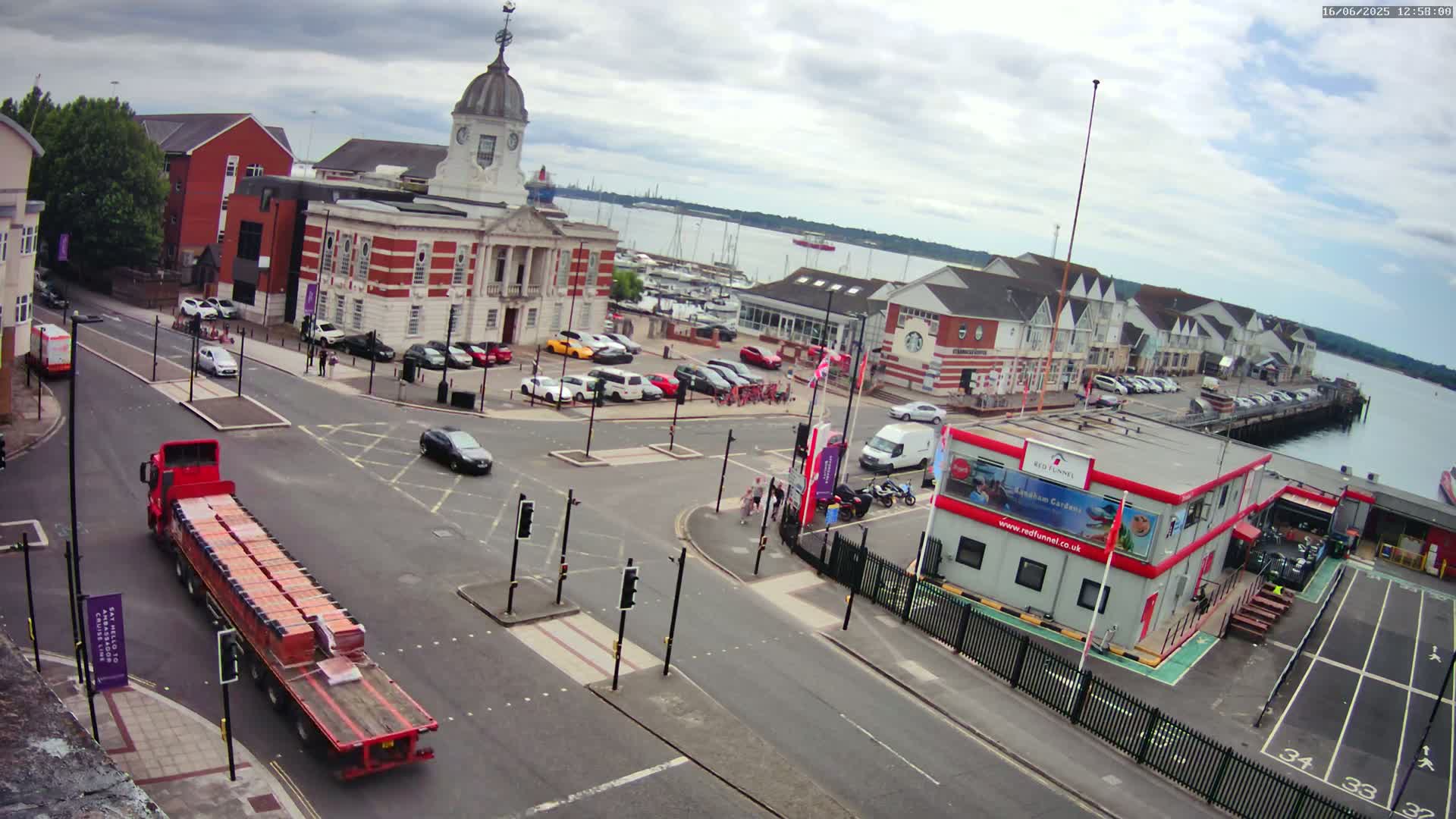 A red truck carrying a long load is driving through a town square near a harbor on an overcast day, with cars parked in lots surrounding a brick building and a waterfront.