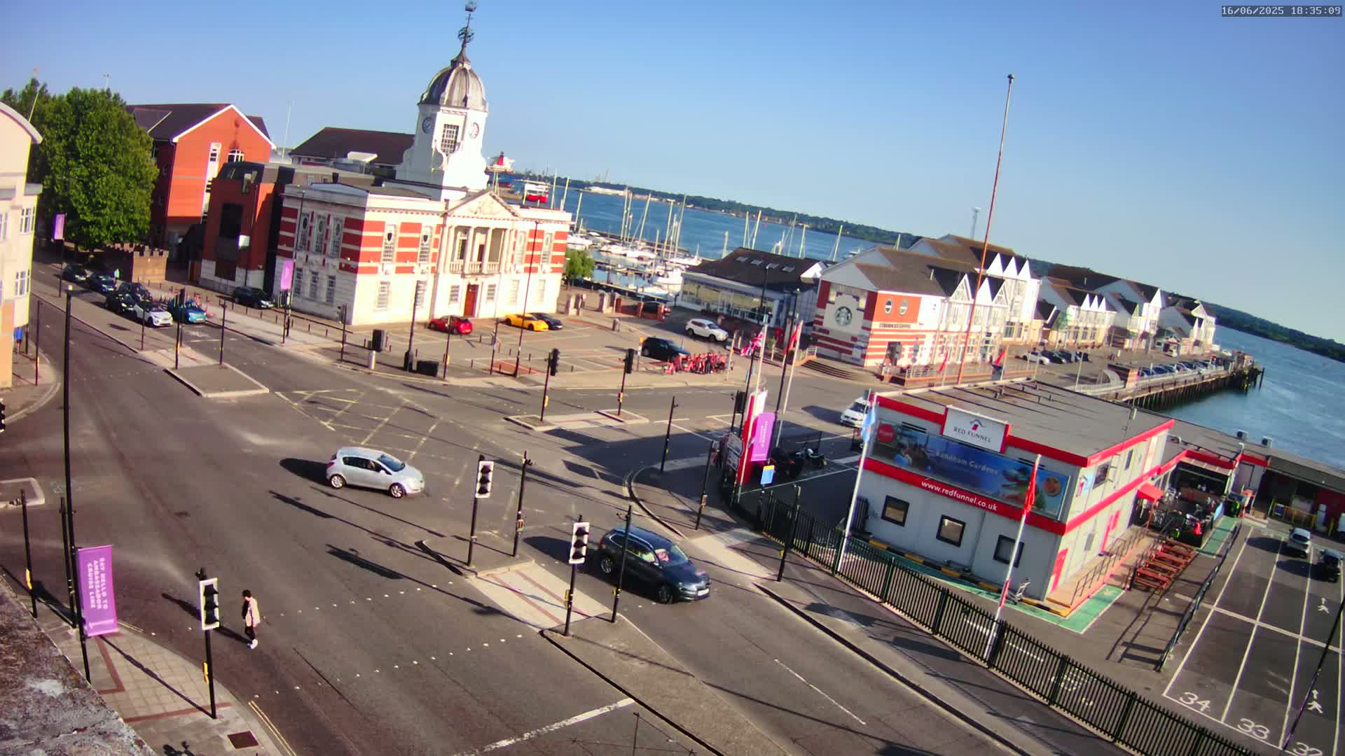A high-angle view of a town square and harbor on a sunny day, with several cars at a traffic intersection near a brick building and a marina filled with boats.
