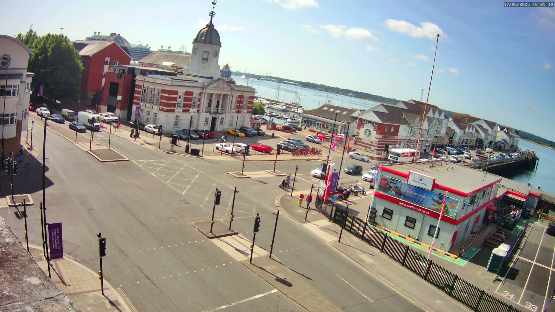 A sunny day overlooking a town square with a brick building, cars parked in a lot, a harbor with boats, and a ferry terminal.