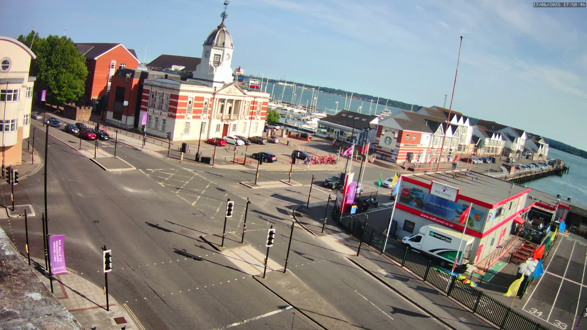 A high-angle, sunny view of a harbor town's intersection with a brick building and a marina, featuring several cars and boats.