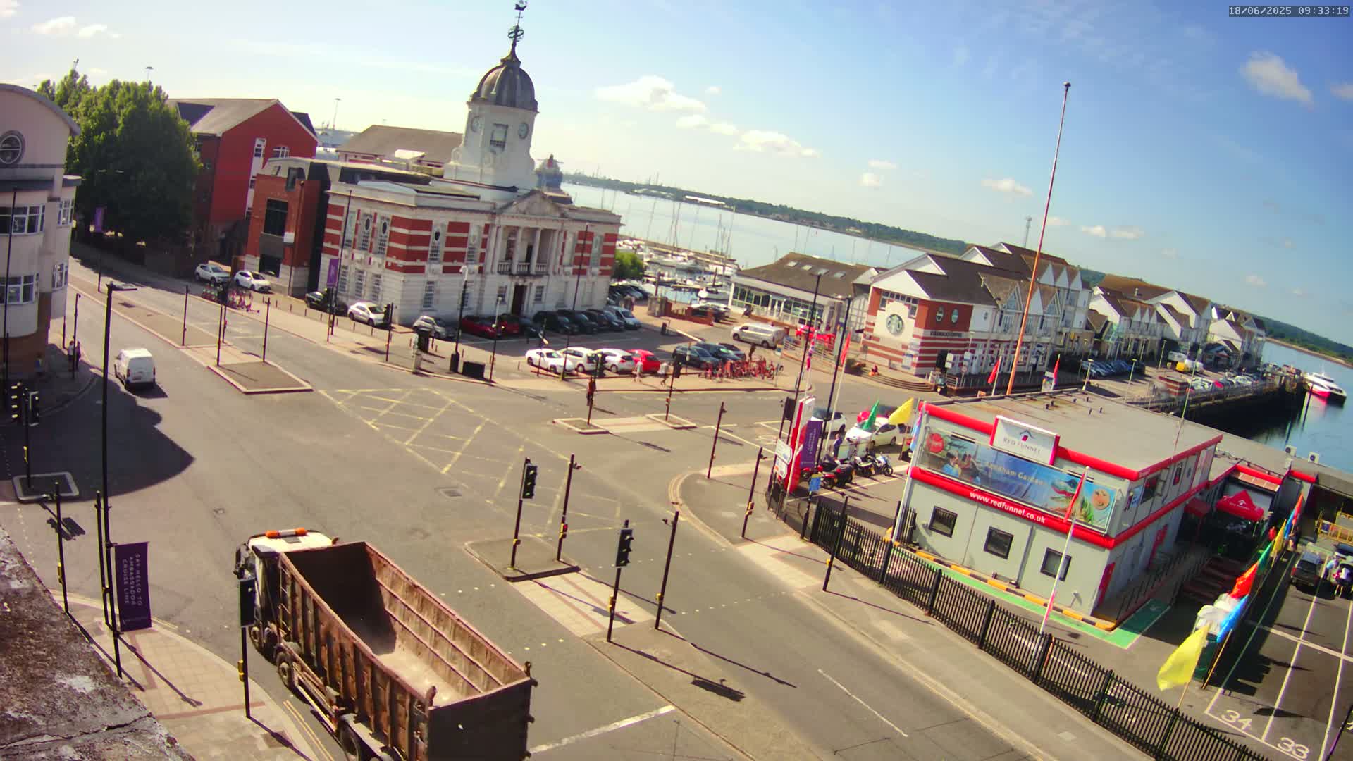A sunny day reveals a harbor town square with a brick building, a parking lot filled with cars, a road intersection, and a waterfront with boats, under a mostly clear sky.