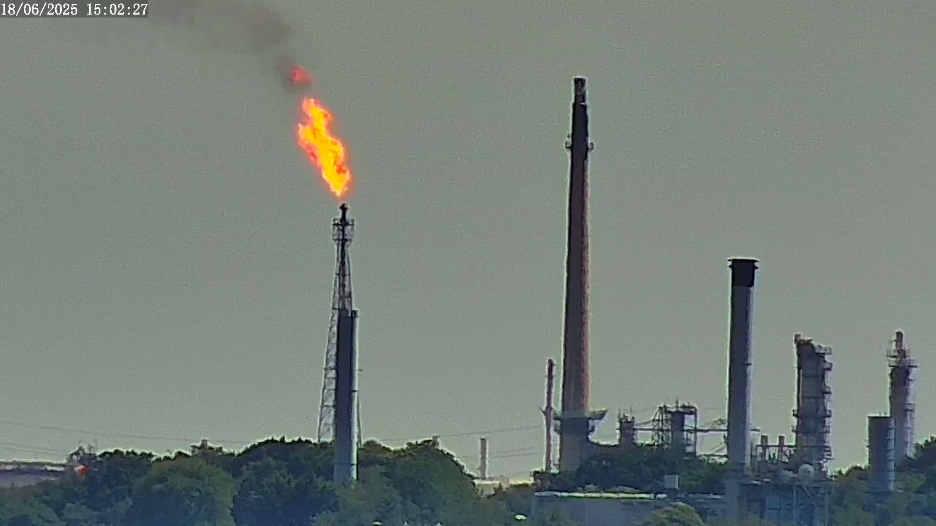 A large industrial plant with tall smokestacks, one of which is burning with a bright orange flame, is visible against a muted, overcast sky.