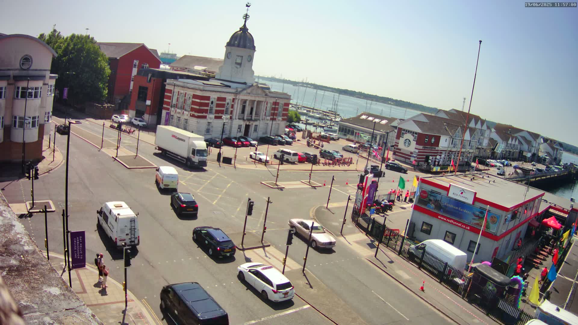 A sunny day overlooking a harbor town square with several vehicles on the roads and a large building in the center.