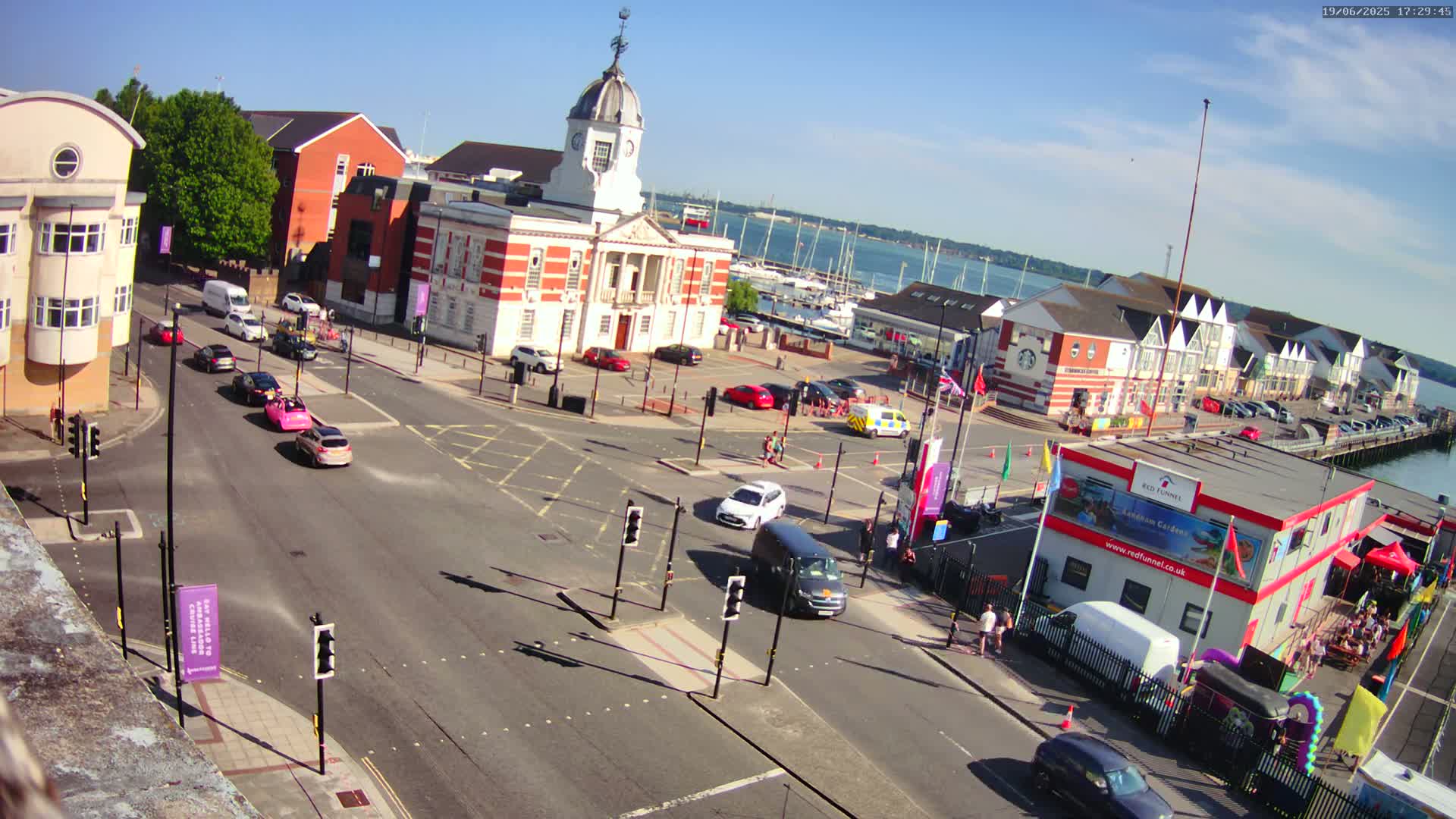 A high-angle, sunny view of a town square near a harbor, with several cars at a crossroads and boats in the water.
