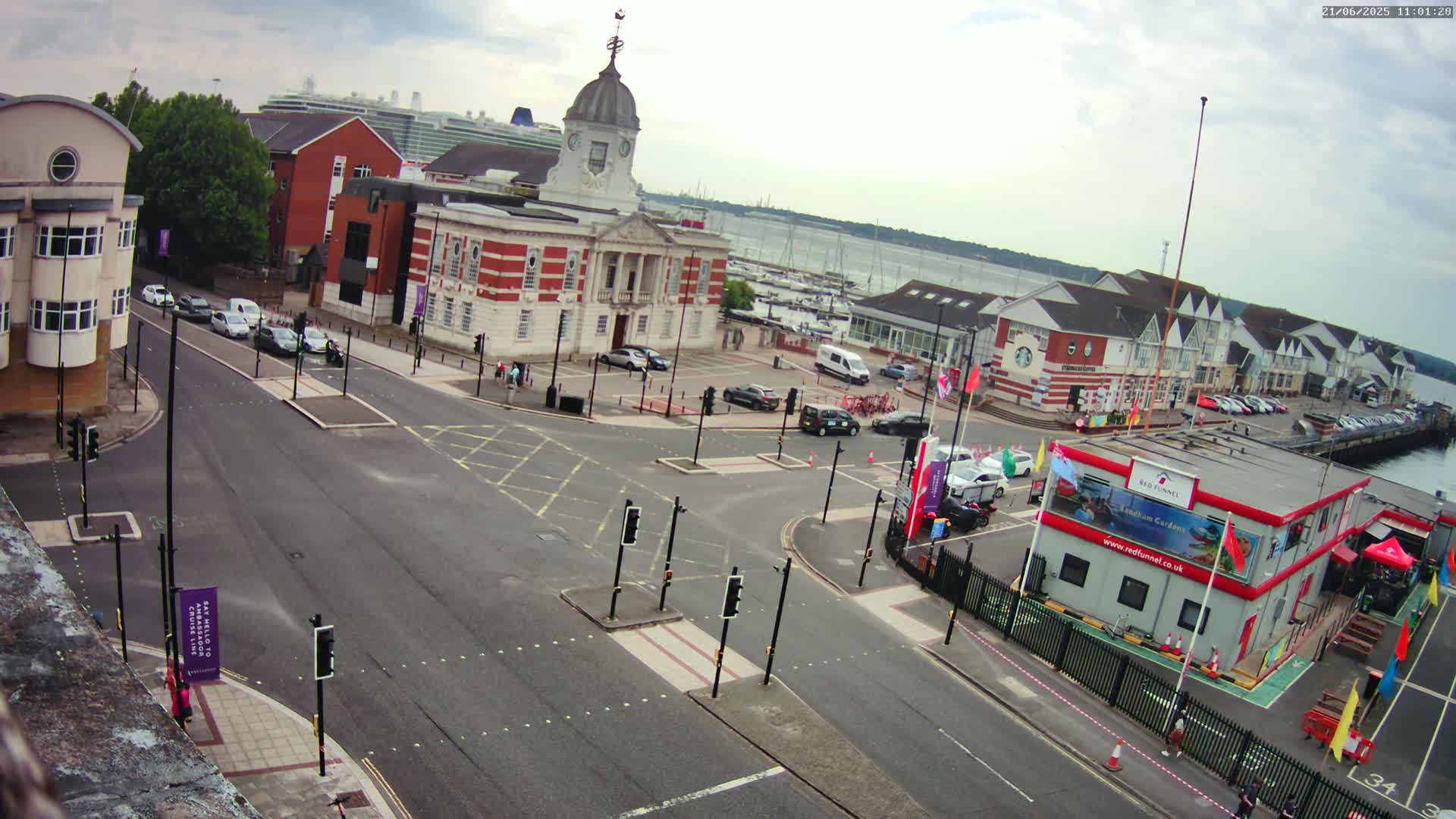 An overcast day reveals a harbor town scene with a large brick building, a cruise ship in the distance, several cars in a parking lot and on roads, and various smaller buildings and boats.