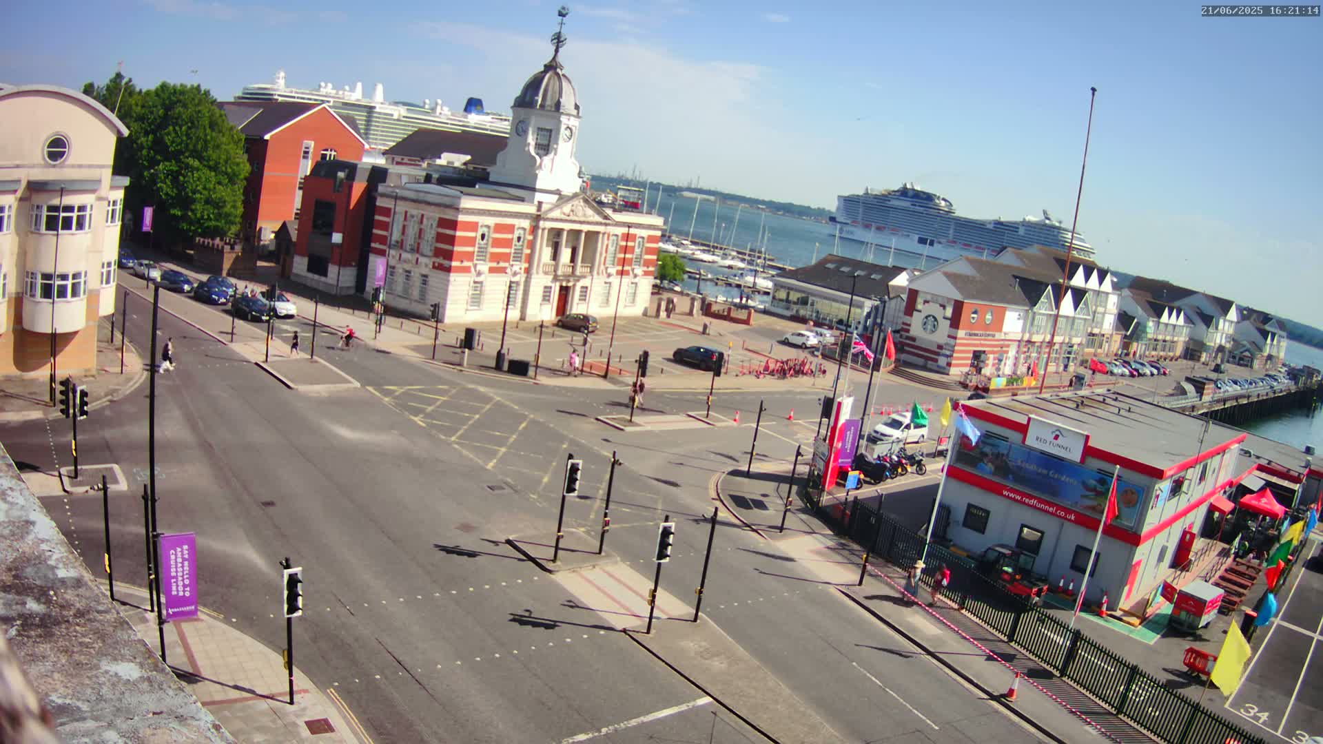 A sunny, high-angle view of a town square with a brick building, intersecting roads, parked cars, and two cruise ships visible in the harbor.