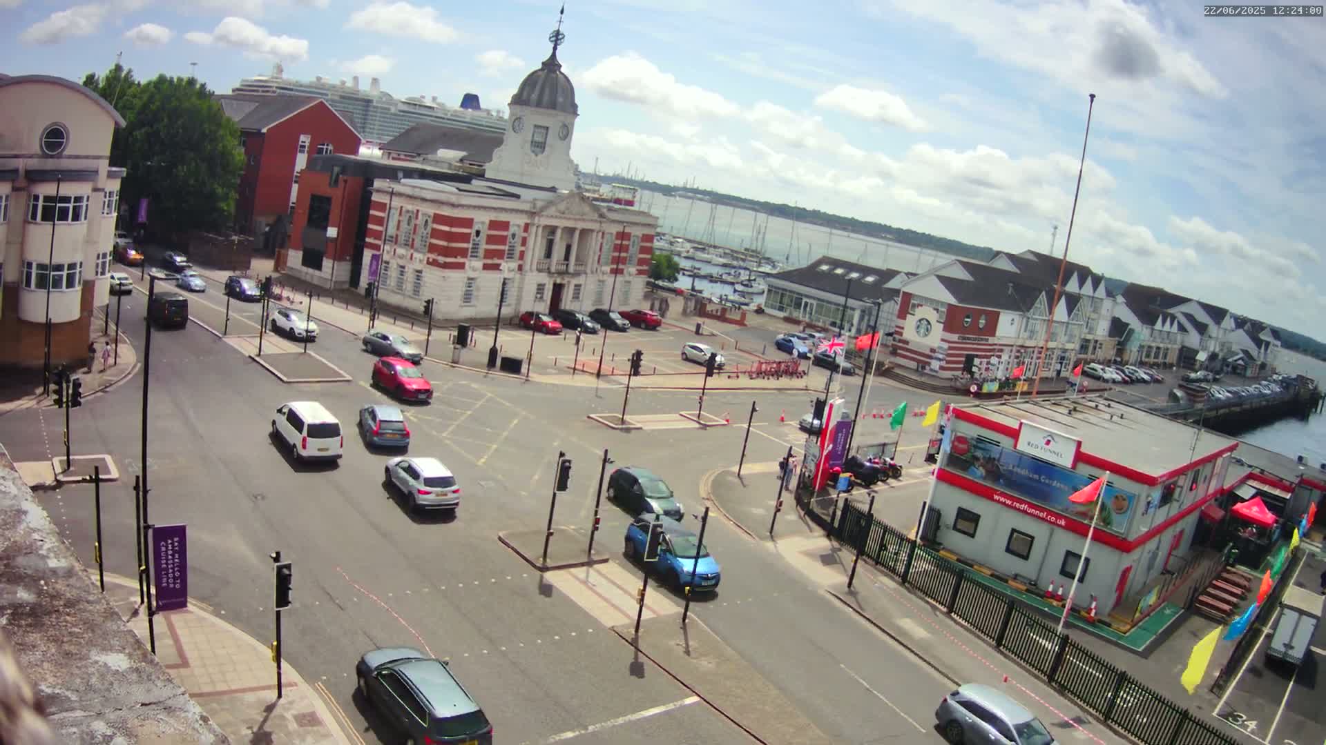 A high-angle view of a town square with several cars at an intersection, a large brick building with a dome, and a cruise ship visible in the background on a partly cloudy day.