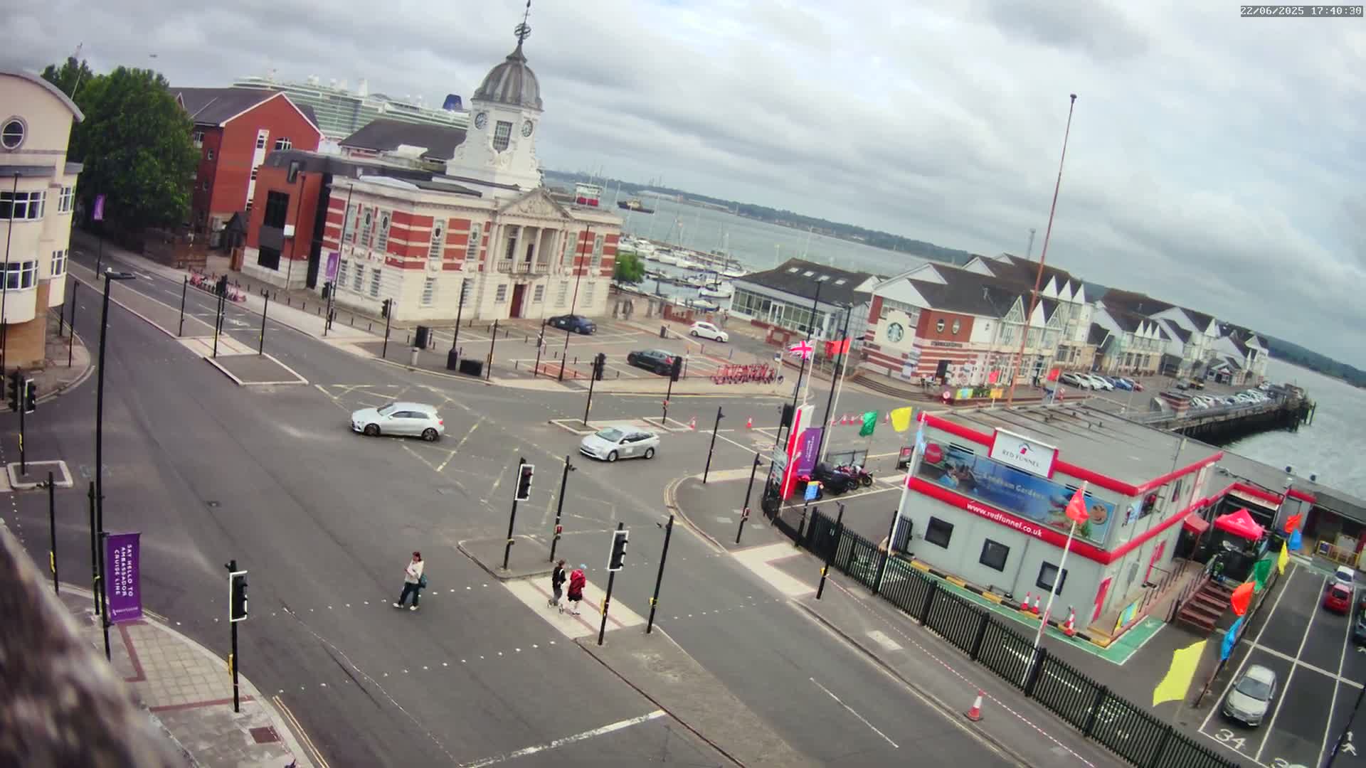 A high-angle view of a town square with a harbor in the background on an overcast day, showing cars driving and pedestrians crossing a street.
