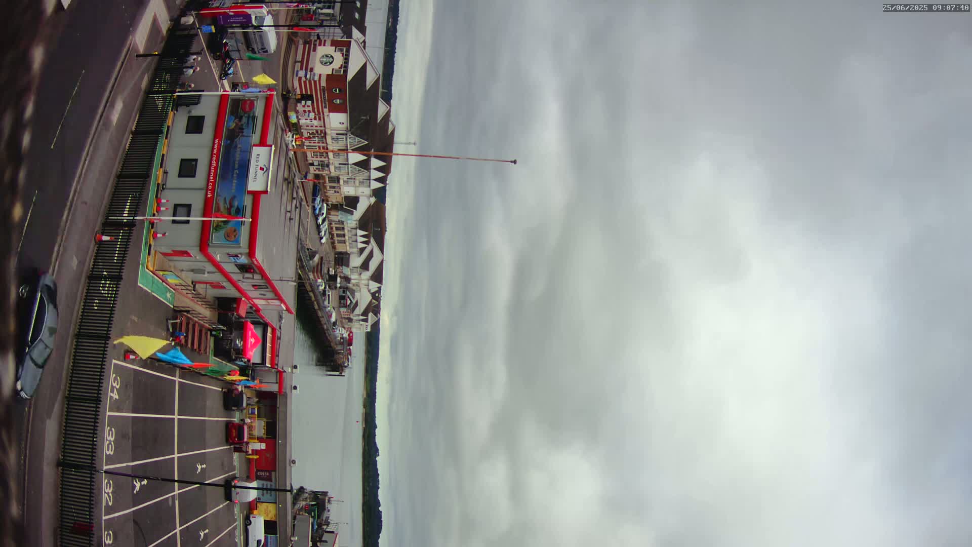 A mostly cloudy sky overlooks a waterfront area with buildings, a parking lot, and a body of water.