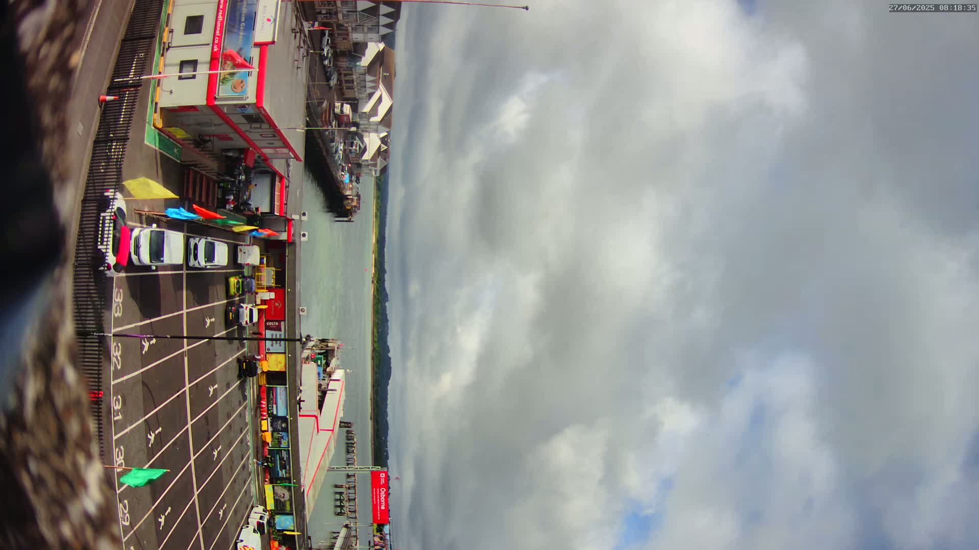 A mostly cloudy sky overlooks a harbor with cars parked in a numbered lot near buildings and a pier.