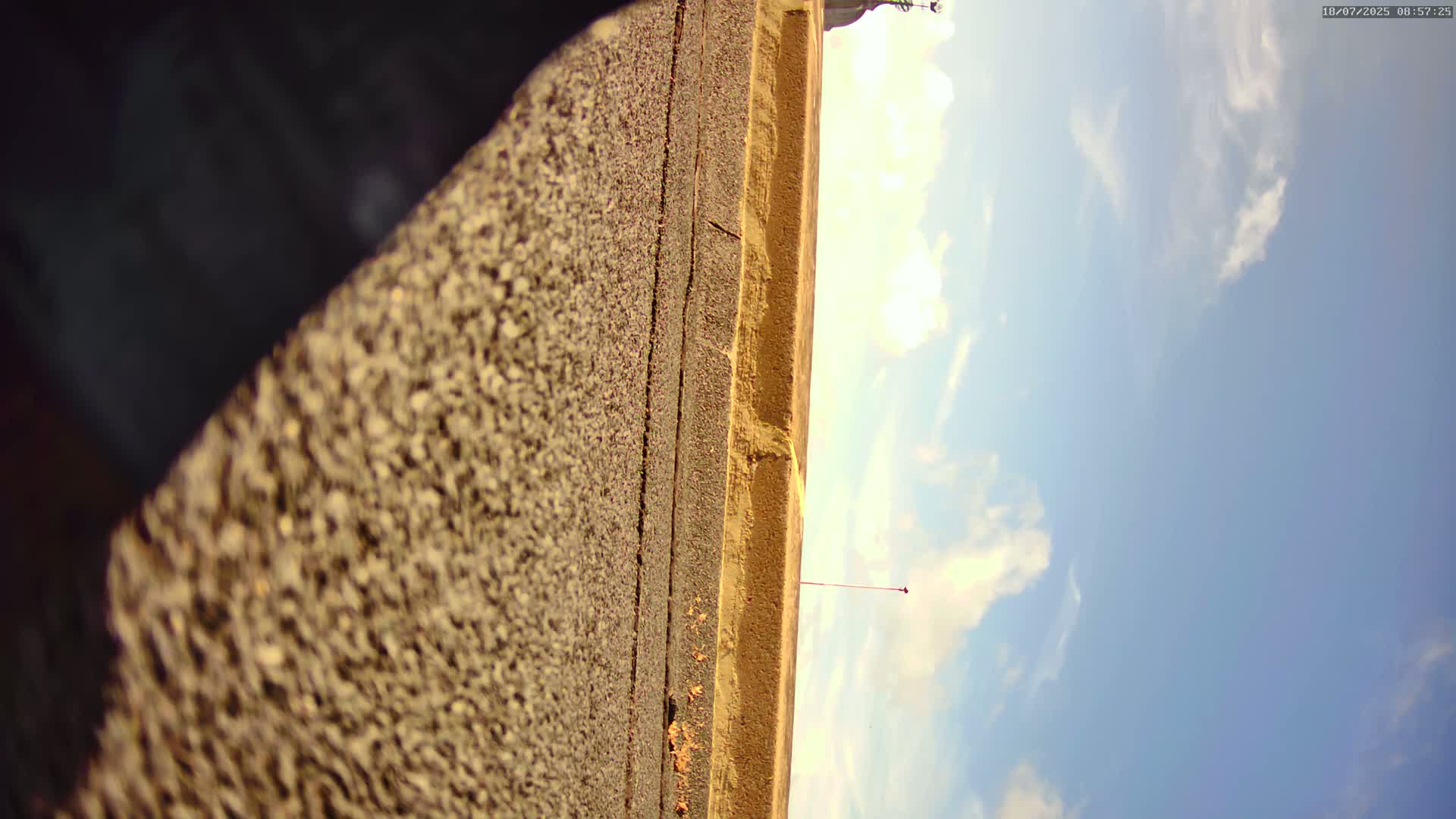 A textured, tan surface angles sharply upward toward a partly cloudy blue sky and a distant, flat, light-brown field.