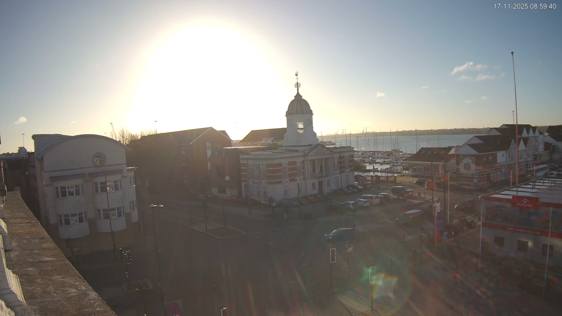 On a bright, sunny day, a historic building with a domed clock tower overlooks a busy waterfront featuring a marina filled with sailboats, adjacent streets with parked cars, and other town buildings.