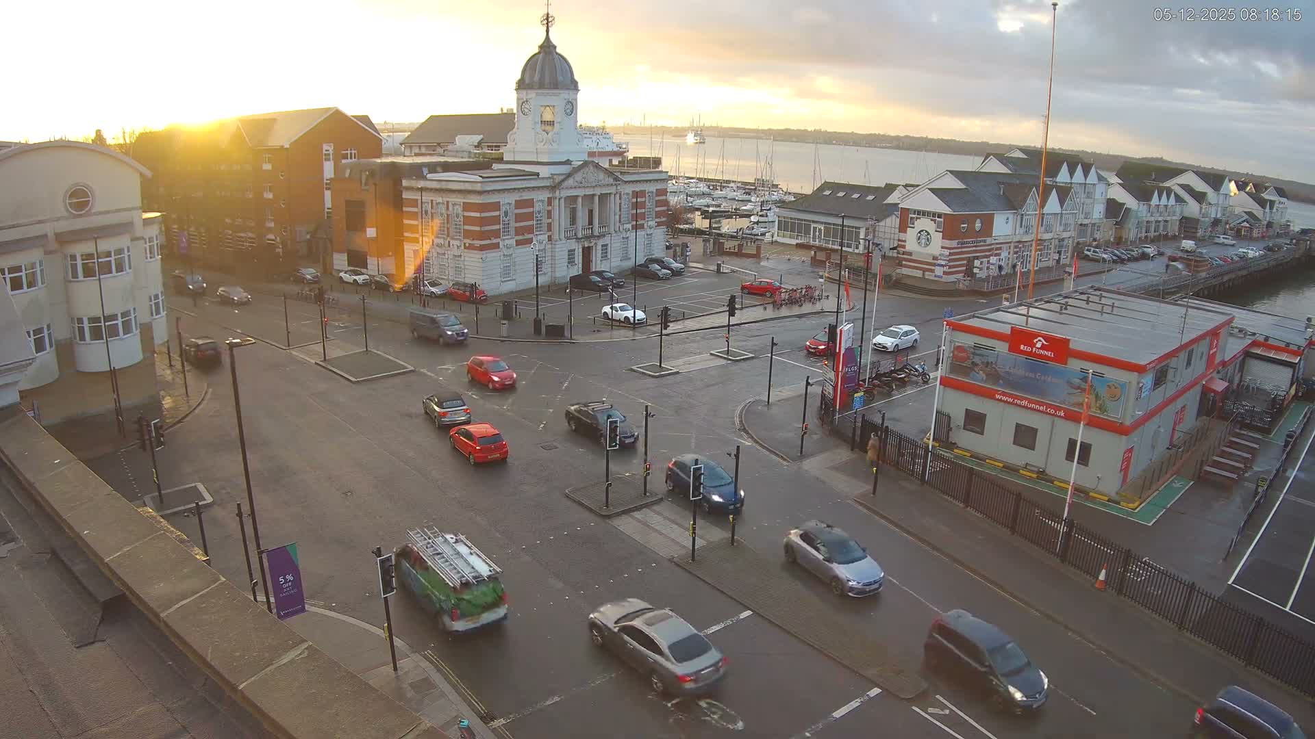 An elevated view reveals a bustling waterfront town with cars driving on wet roads past a historic clock tower building, a marina filled with boats, and a ferry terminal, under the golden light of an early morning sun shining through scattered clouds.