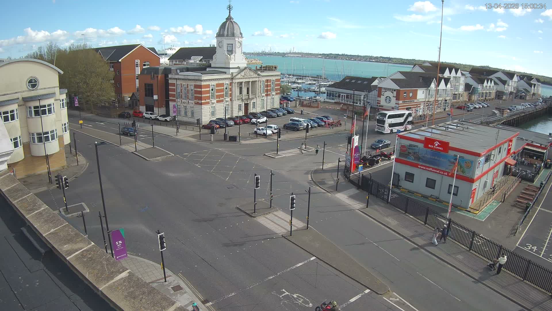 An elevated view reveals a bustling waterfront town with cars driving on wet roads past a historic clock tower building, a marina filled with boats, and a ferry terminal, under the golden light of an early morning sun shining through scattered clouds.