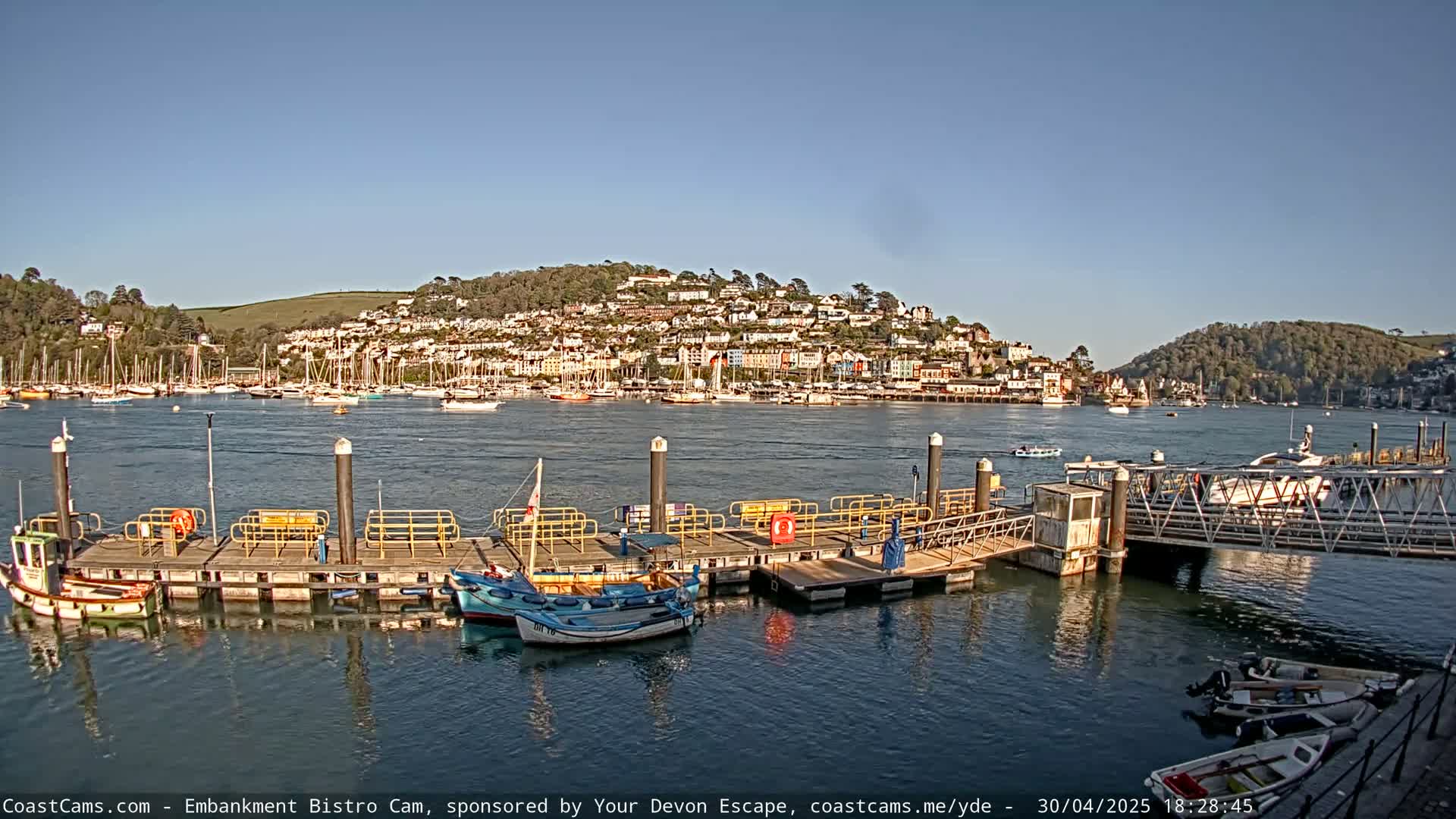 A waterfront scene on a sunny day shows a harbor filled with boats, a dock with small fishing boats, and a hillside town in the background.