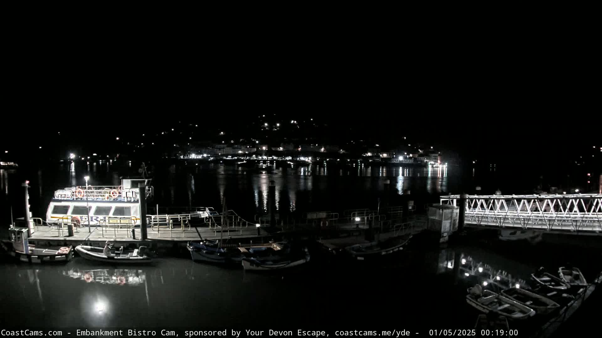 At night, a harbor scene shows several docked boats and a ferry at a pier, with a town's lights reflecting on the calm water in the background.