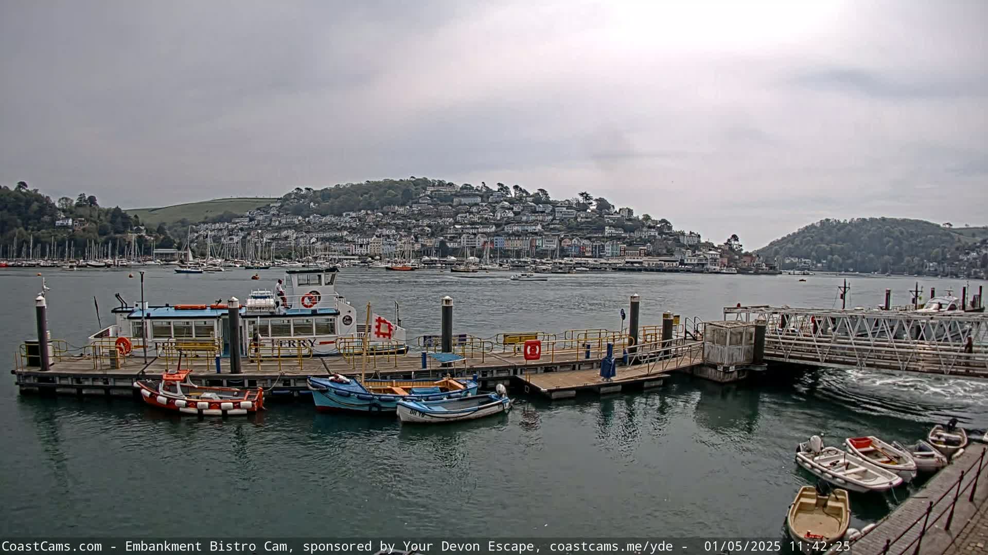 A ferry is docked at a pier in a harbor, with numerous boats and a town nestled on a hillside in the background under an overcast sky.