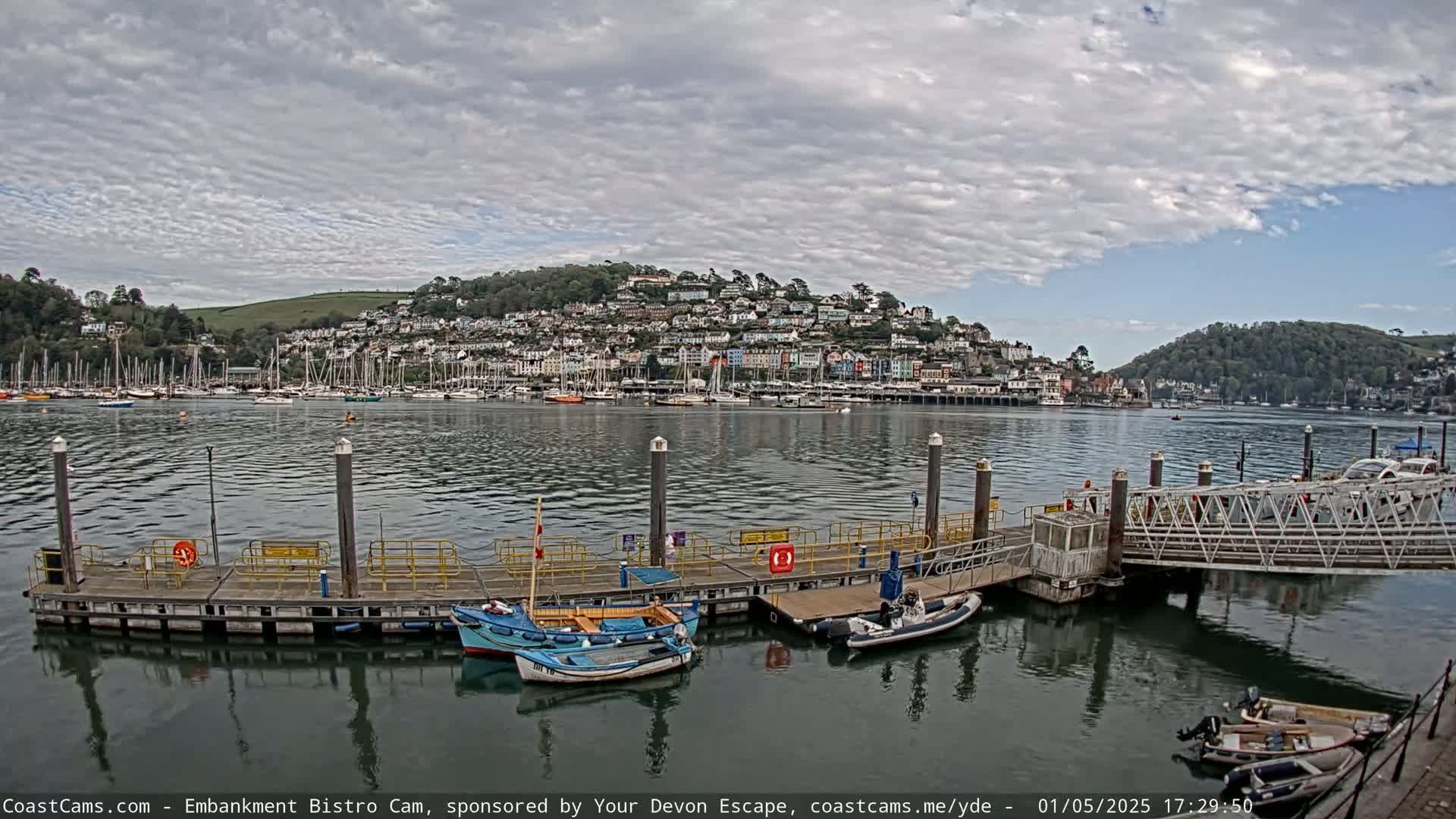 A harbor with several boats docked at a pier, under a partly cloudy sky, overlooks a hillside town.