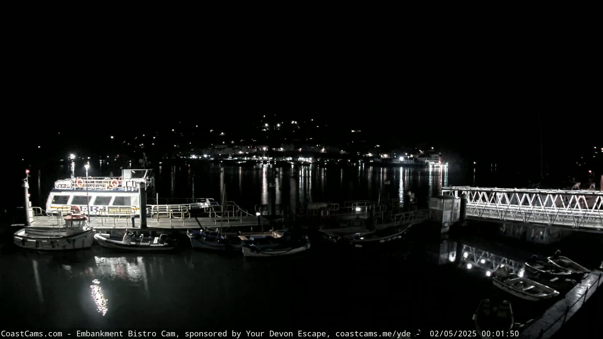 A nighttime harbor scene shows a docked ferry and several smaller boats, with a town's lights reflecting in the calm water.