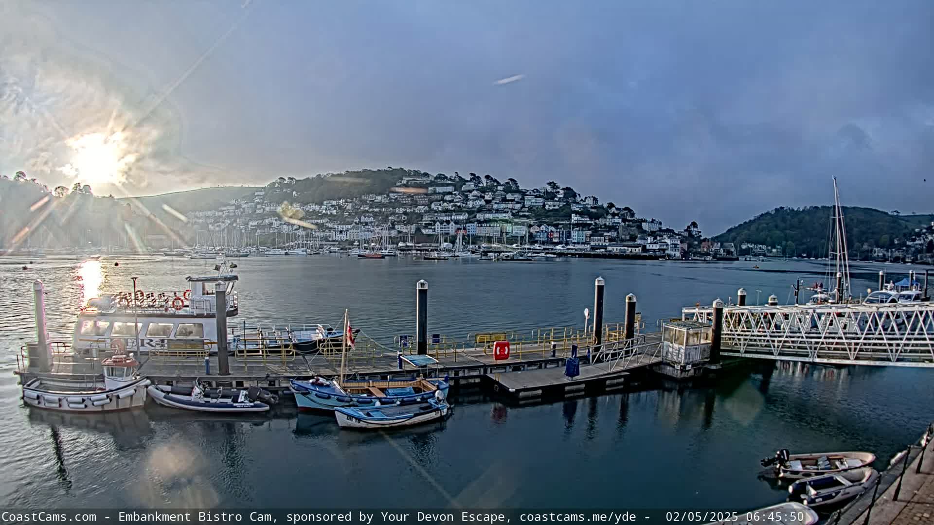 A calm harbor scene at sunrise shows a town nestled on a hill overlooking a dock with several small boats and a larger ferry.