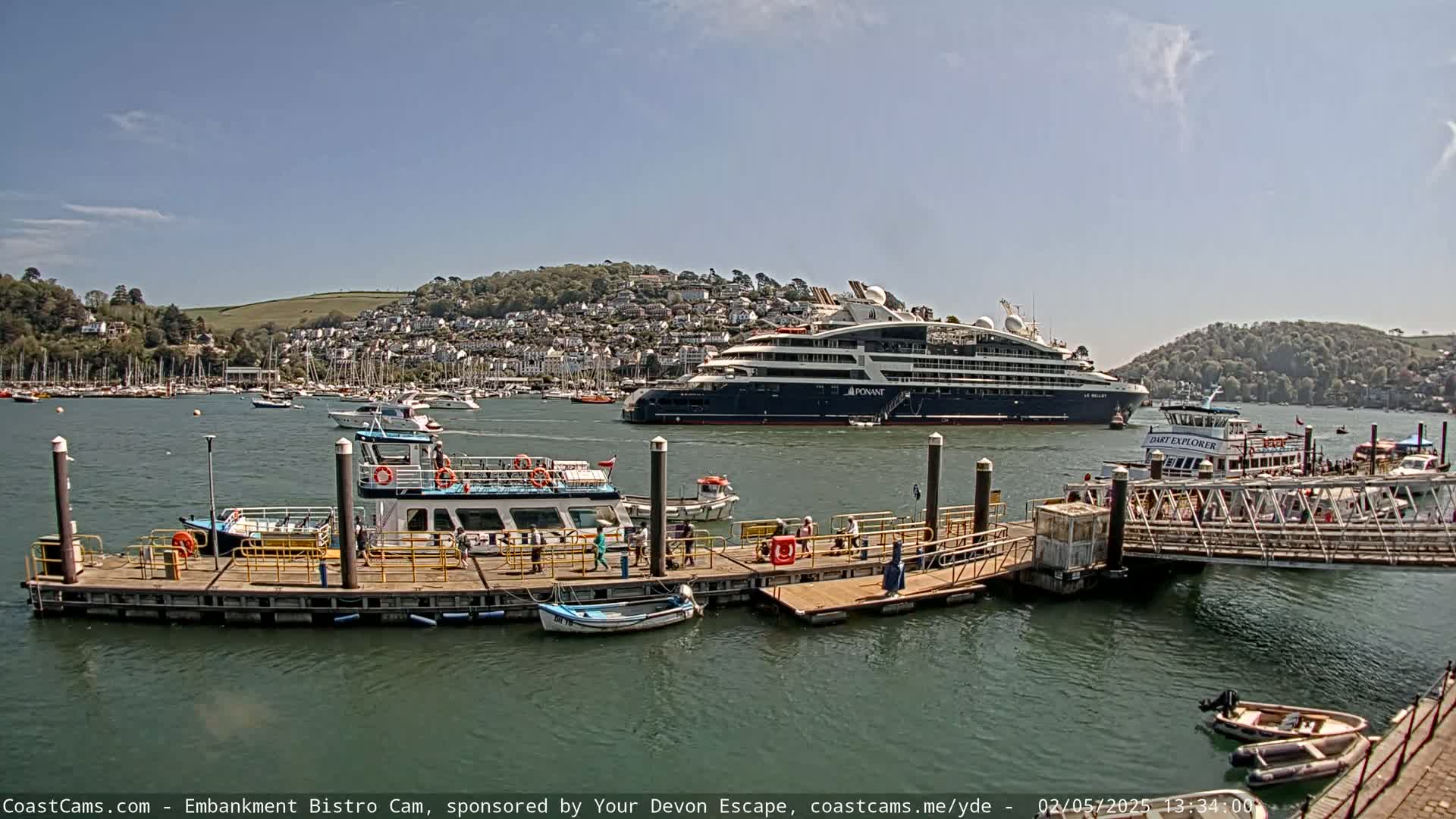 A large cruise ship is moored in a harbor on a sunny day, near a dock where several smaller passenger boats and other vessels are also present.