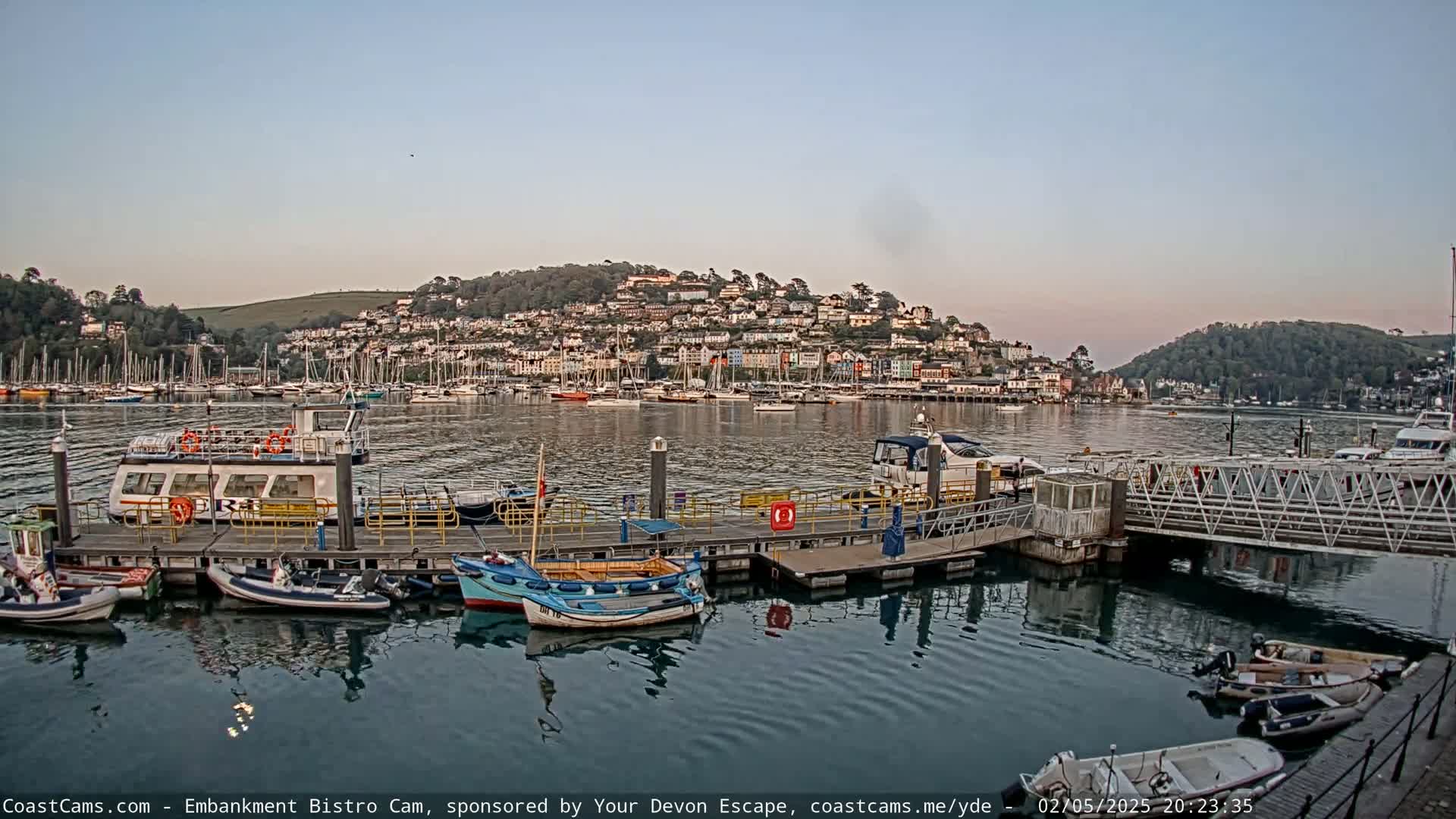 A calm harbor scene at dusk shows a docked ferry, several smaller boats, and a hill covered in colorful houses.