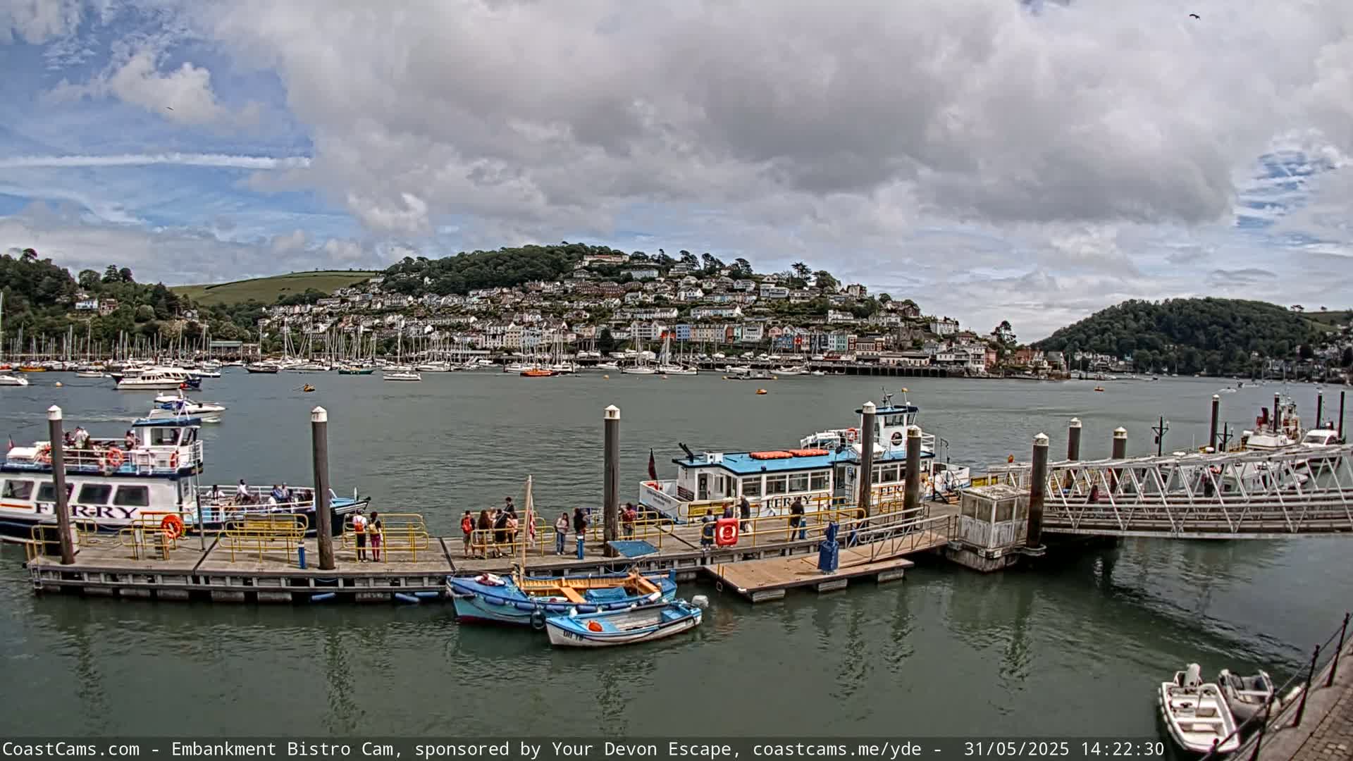 A ferry terminal with several boats docked, some passengers, and a town on a hill in the background under a partly cloudy sky.