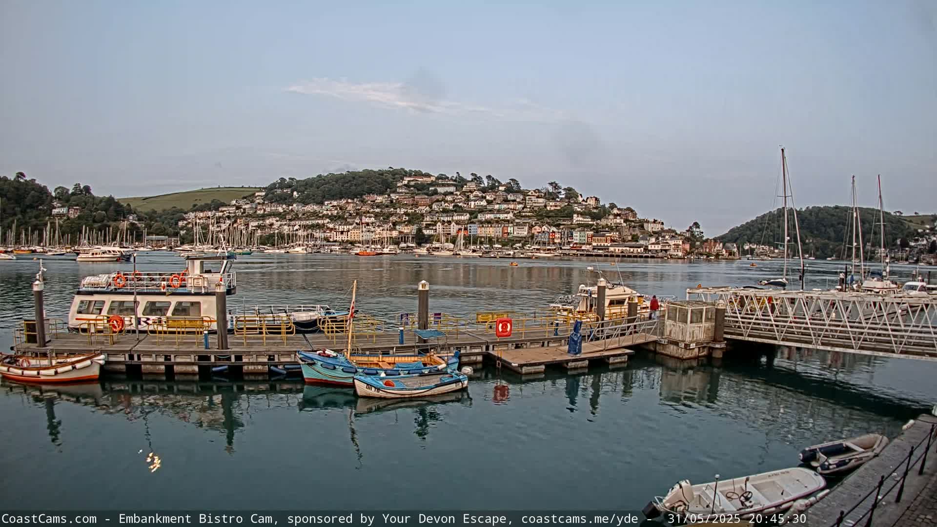 A calm harbor scene shows several boats moored at a dock, with a hillside town visible in the background under a partly cloudy sky.