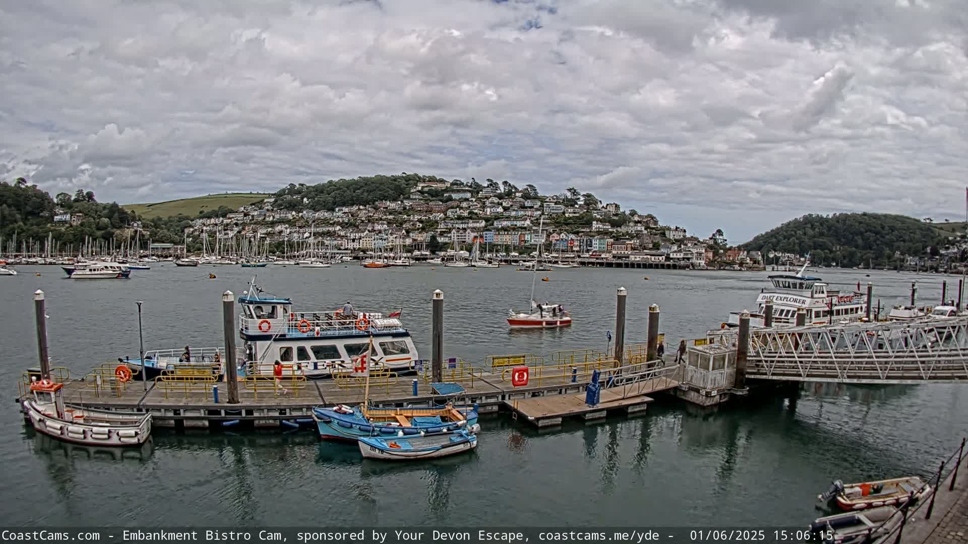A harbor scene on an overcast day shows several boats docked at a pier, with a town nestled on a hillside in the background.