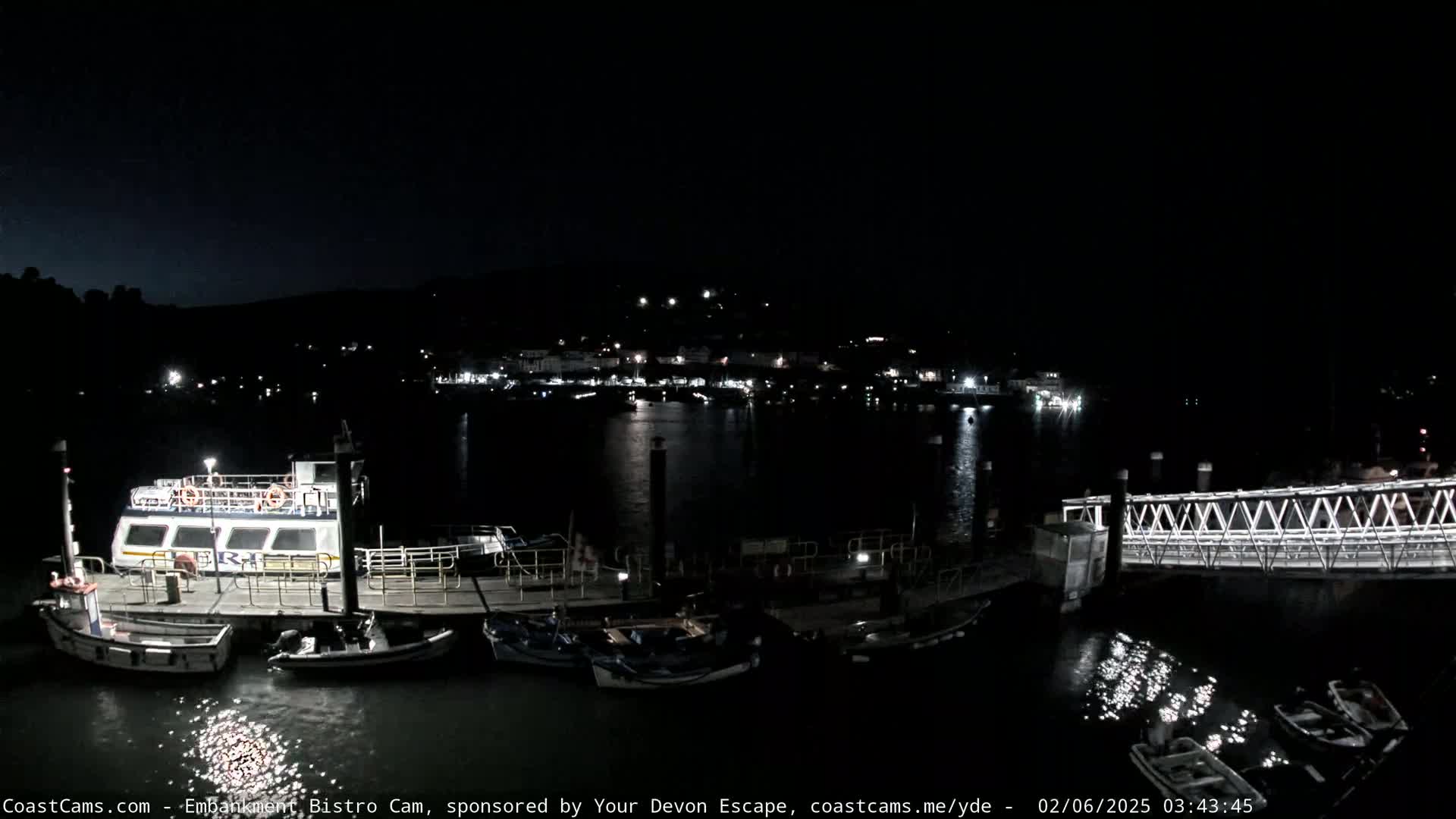 A nighttime view of a harbor with several boats docked at a pier, a larger boat moored alongside, and a small bridge extending across the water, all under a clear, dark sky.