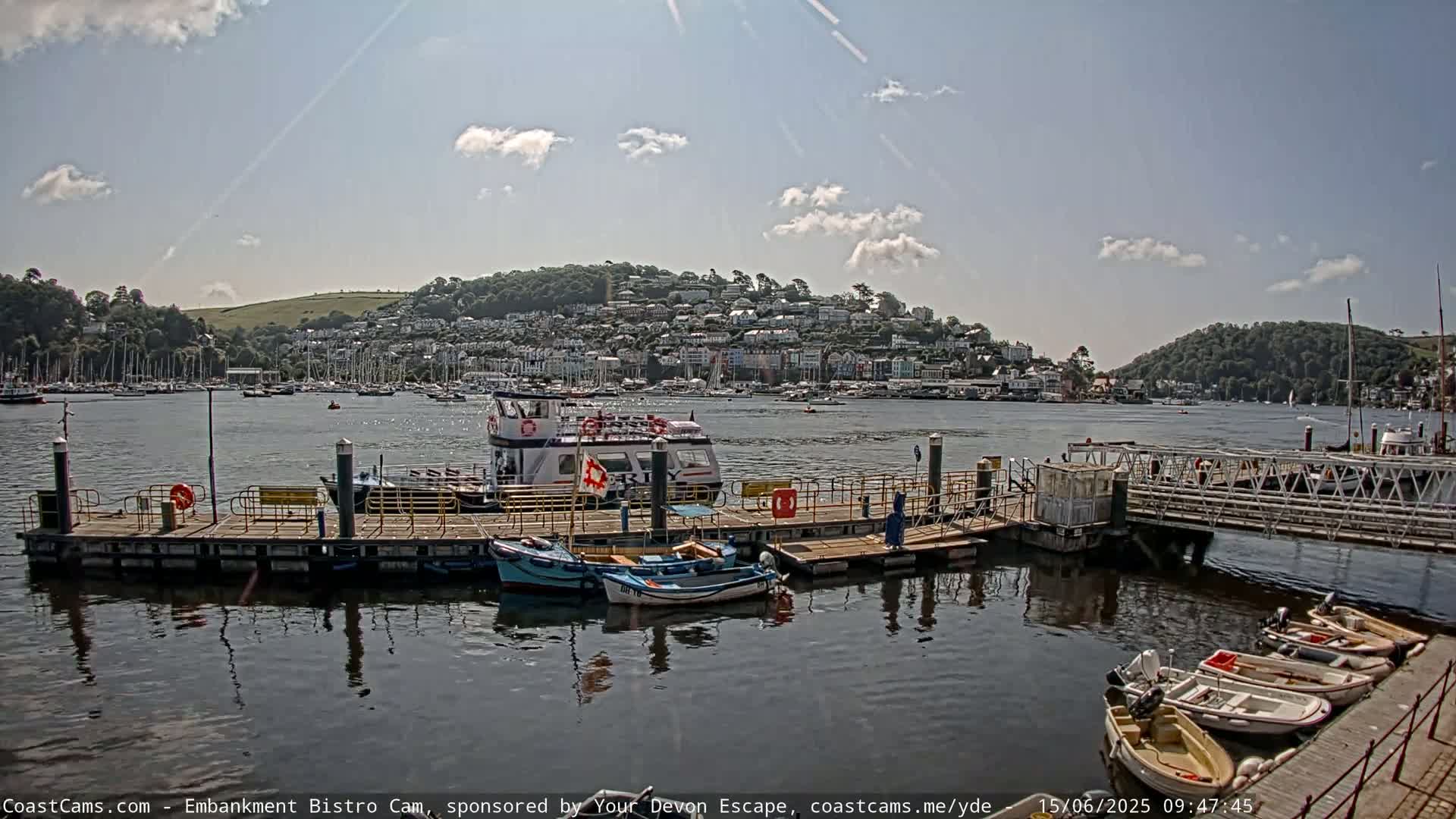 A sunny day shows a harbor with a docked ferry, several smaller boats, and a town nestled on a hillside in the background.