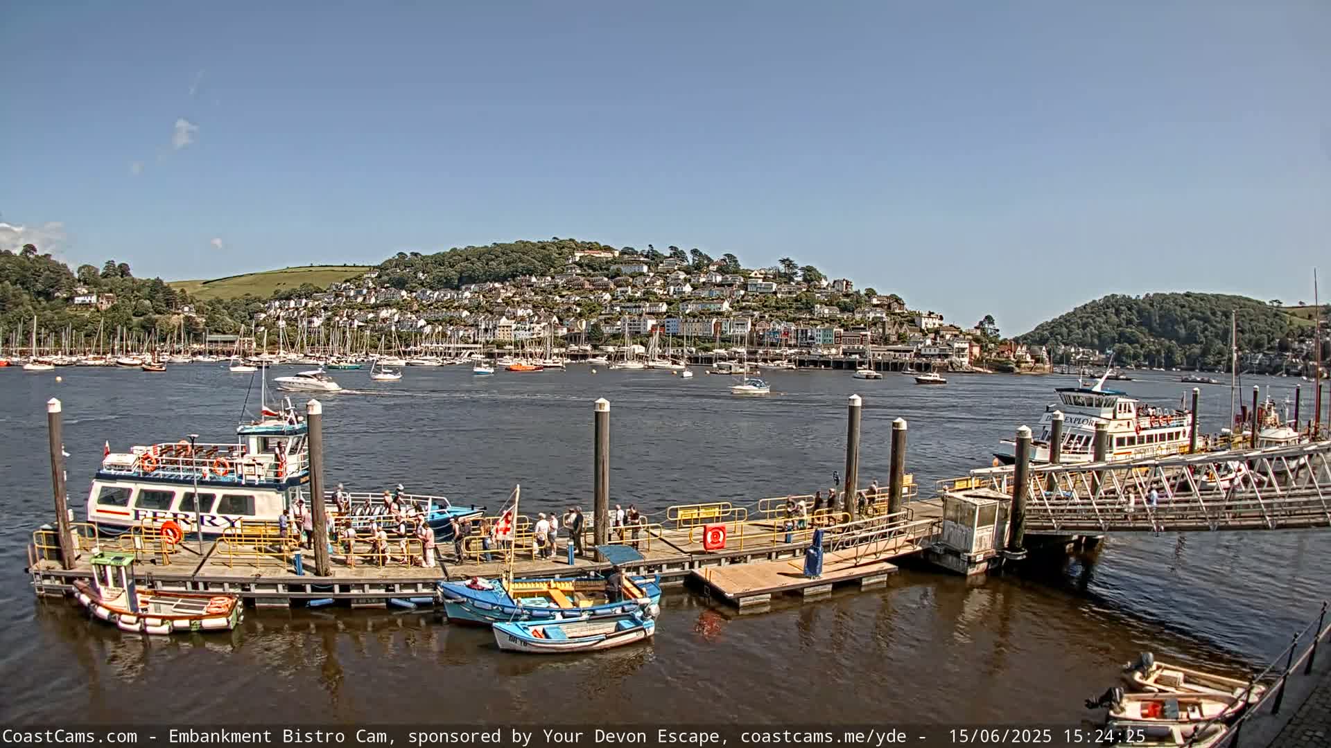 A ferry is docked at a pier with several smaller boats, and a town sits on a hill across a calm body of water under a clear blue sky.