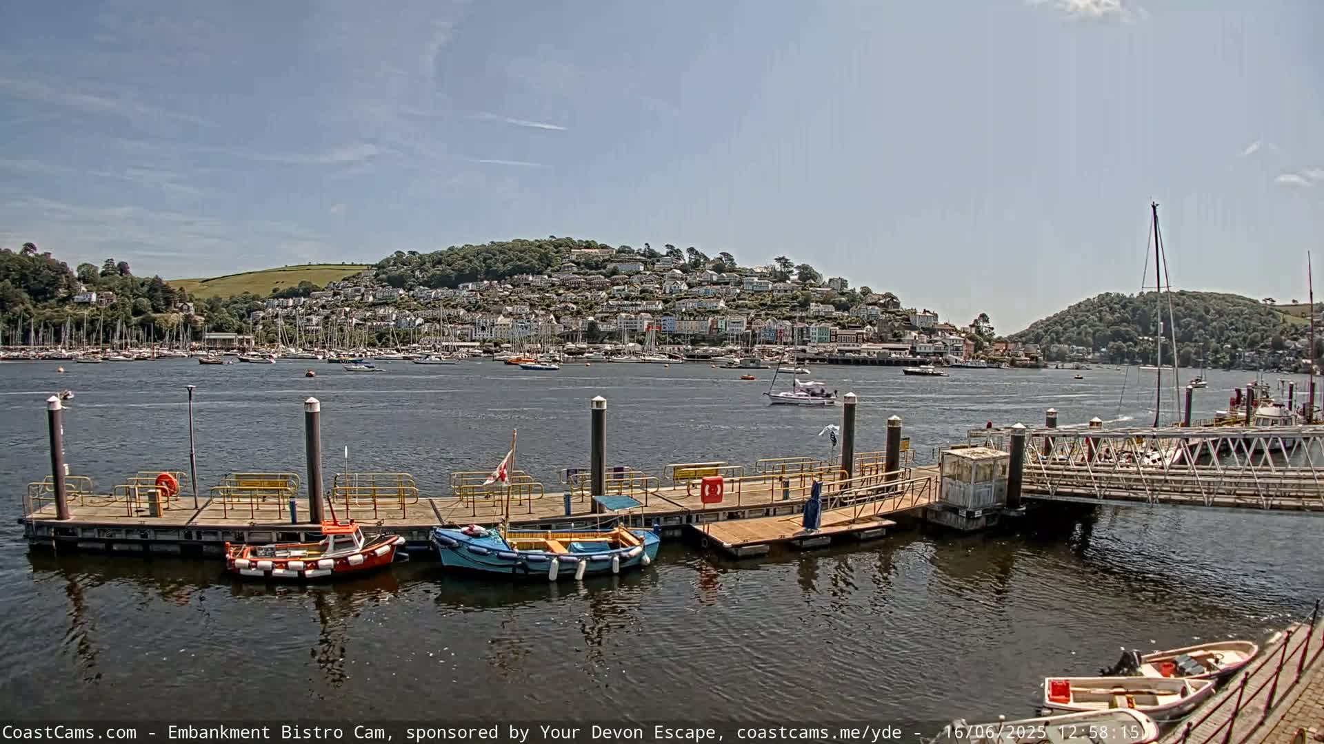 A harbor with several small boats moored at a dock, overlooking a town nestled on a hillside under a mostly sunny sky.