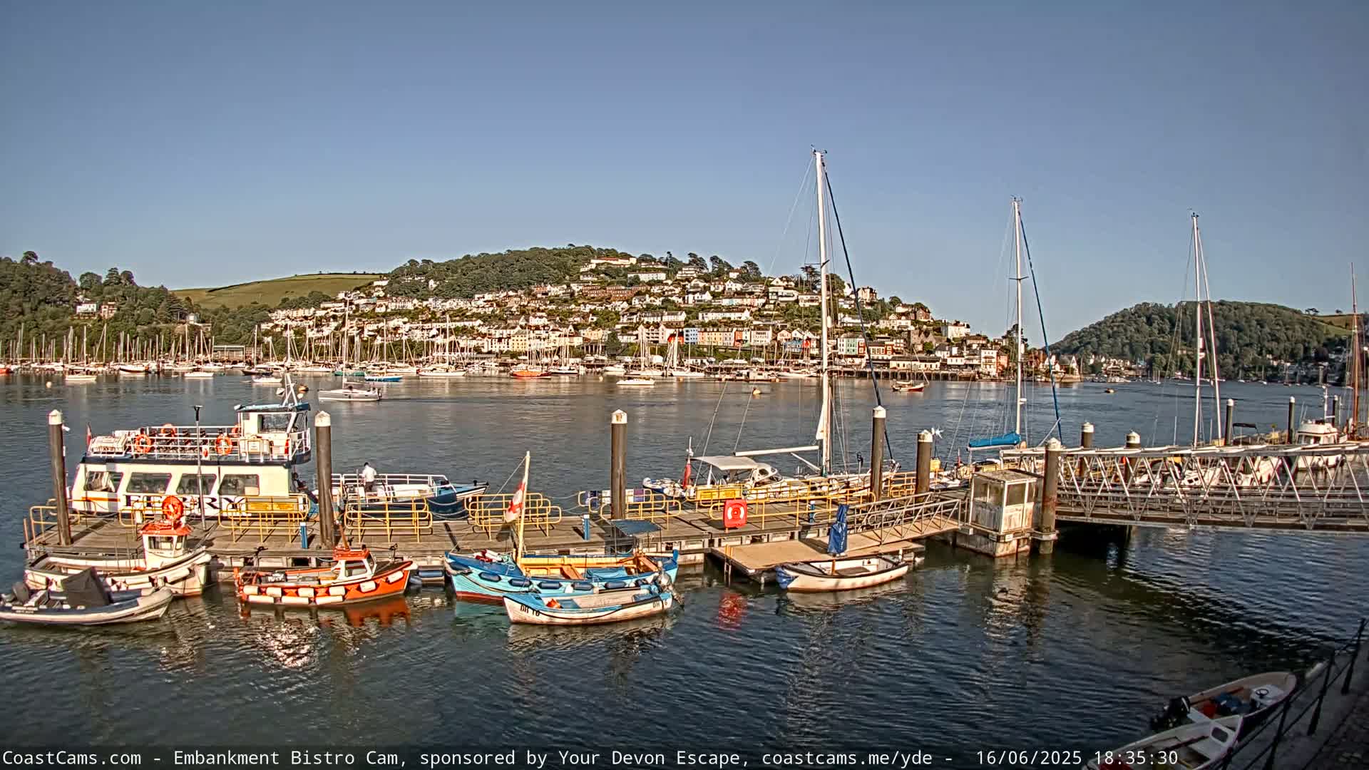 A sunny day shows a harbor filled with boats and a hilltop town in the background.