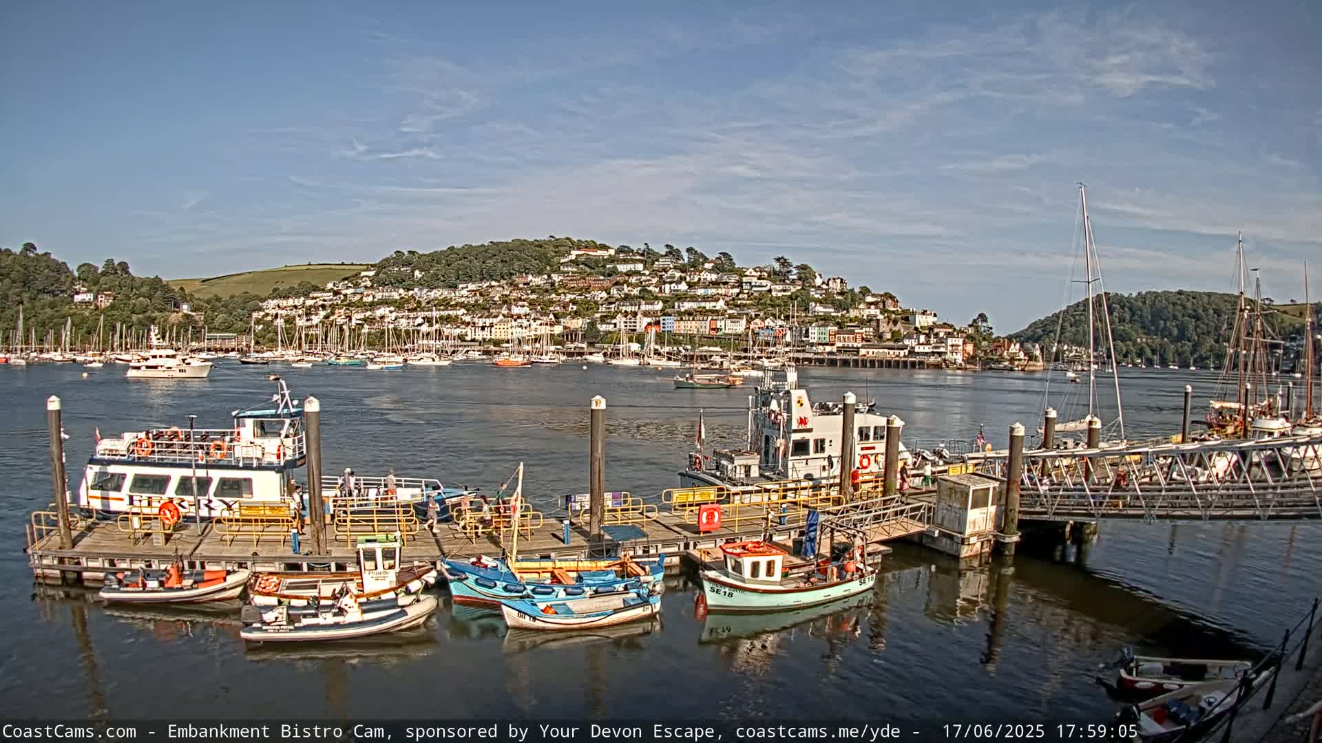 A harbor scene on a sunny day shows a ferry docked at a pier with several smaller boats, a town nestled on a hillside in the background.